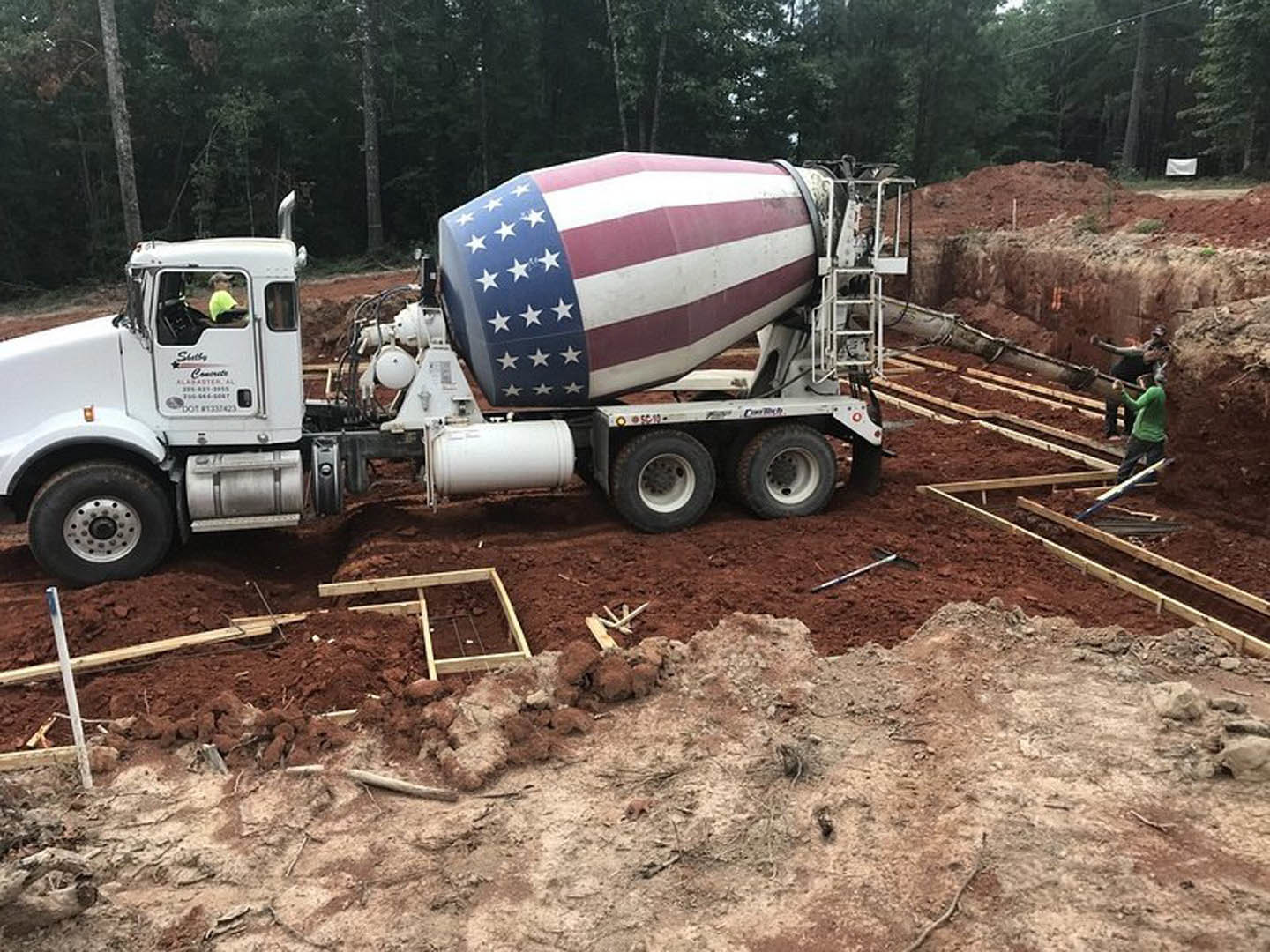 Concrete mixer truck parked outdoors with American flag attached, surrounded by trees and ground, visible wheels and tires
