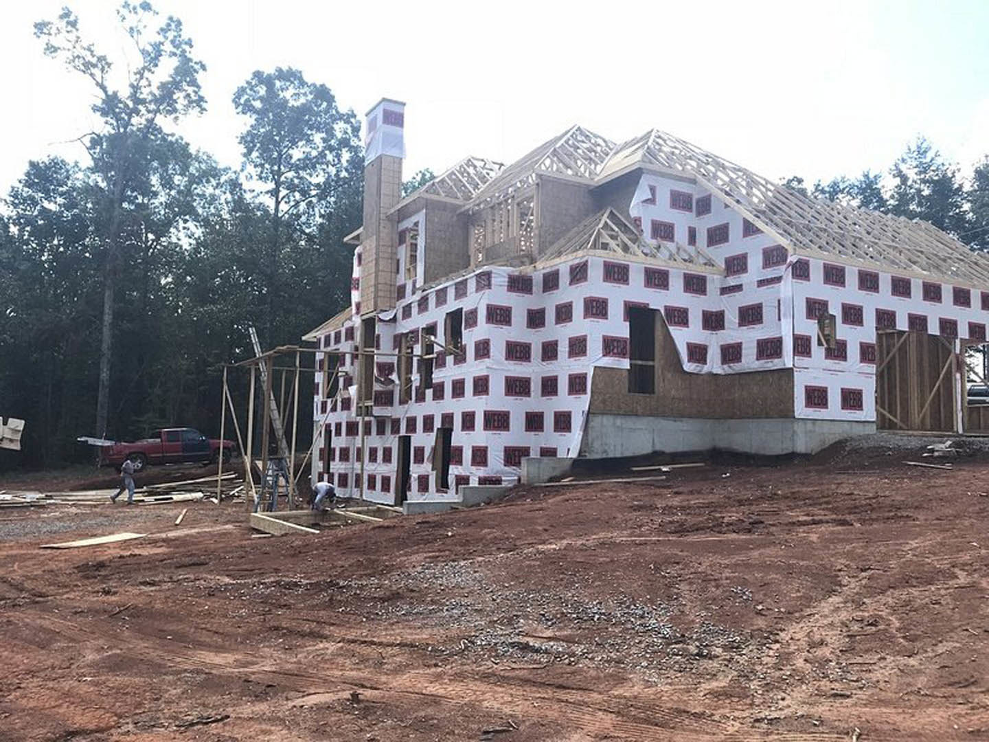 Partially built house with red and white square panels, exposed framing, white protective covering, dirt lot, red truck parked nearby, trees in background
