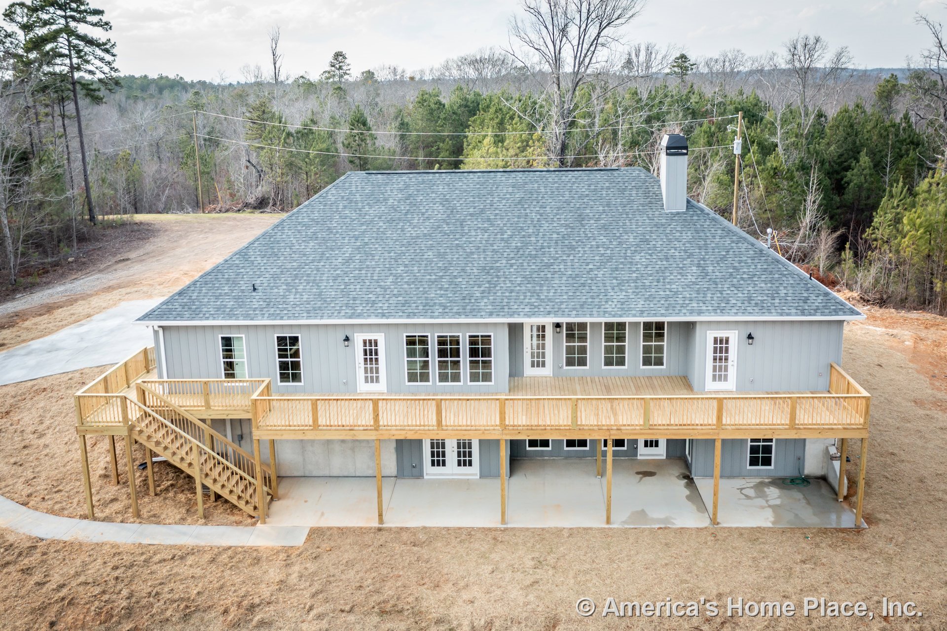 Large elevated wooden deck with stairs, multiple exterior doors, gray vertical siding, double-hung windows, concrete patio beneath deck, asphalt shingle roof, exterior lighting
