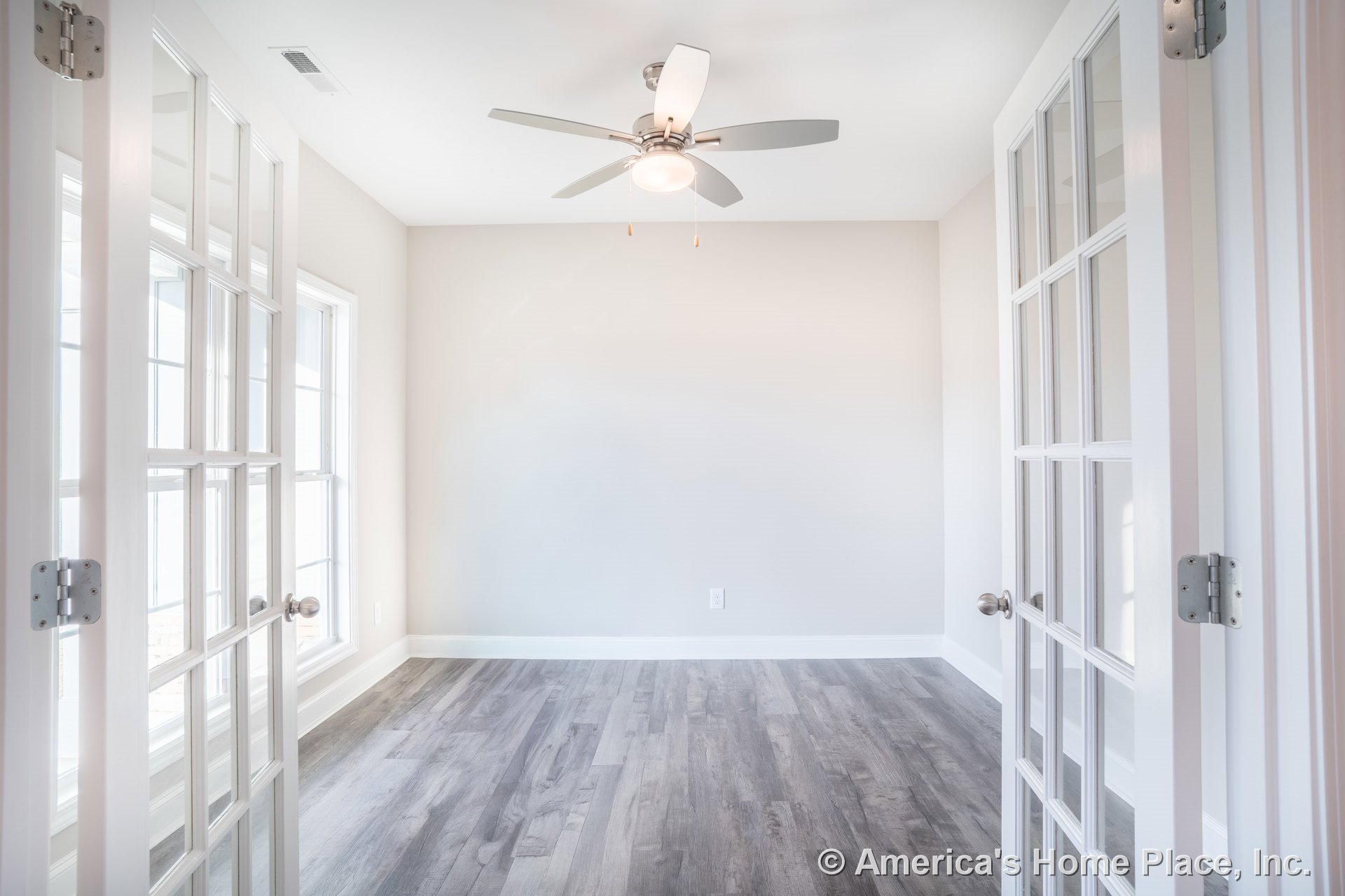 Double glass French doors and large windows with white trim fill the room with natural light; modern ceiling fan with integrated light fixture hangs above gray wood-look flooring