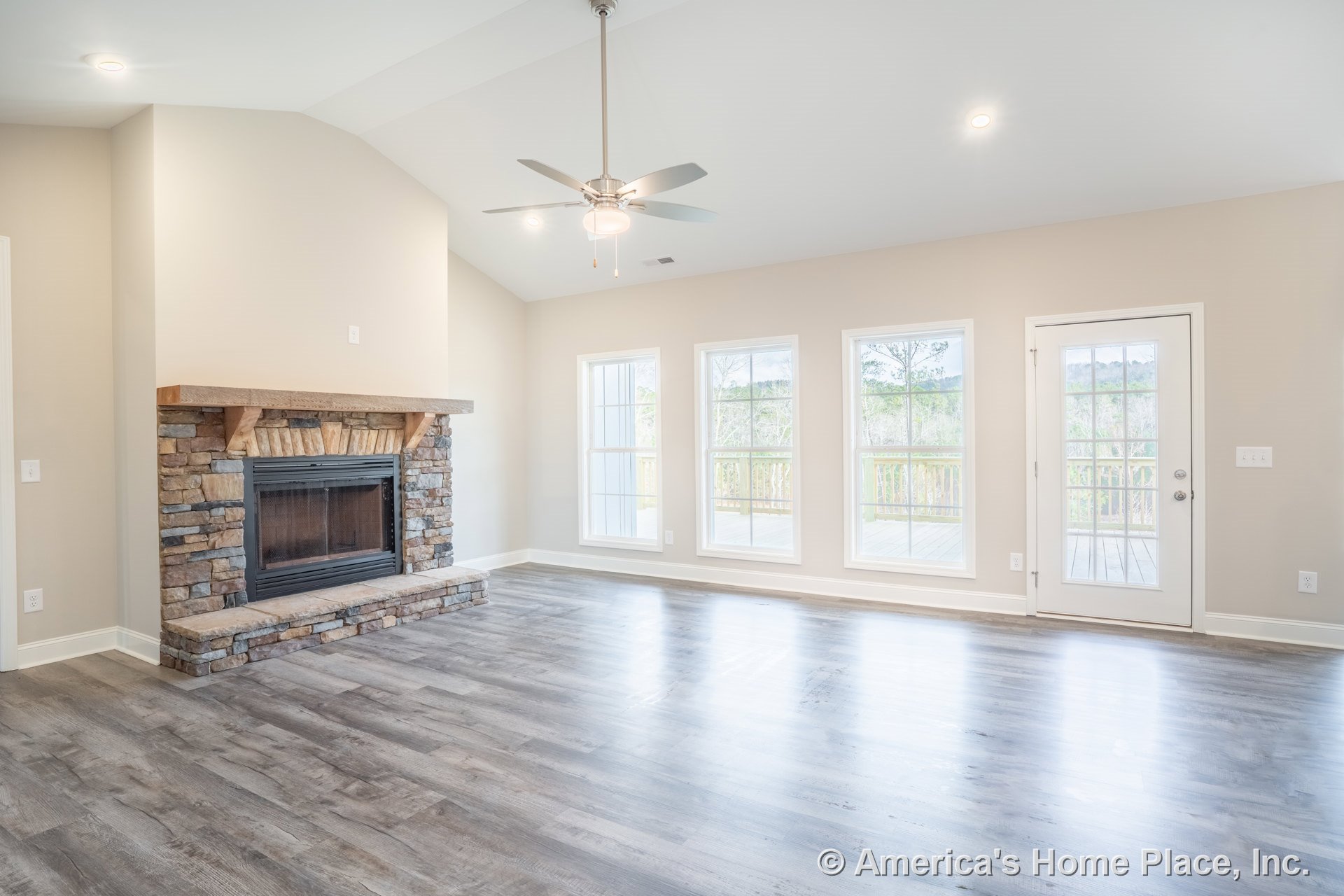 Stone fireplace with wood mantel centered beneath vaulted ceiling featuring recessed lighting and ceiling fan; three large windows and glass exterior door provide porch access