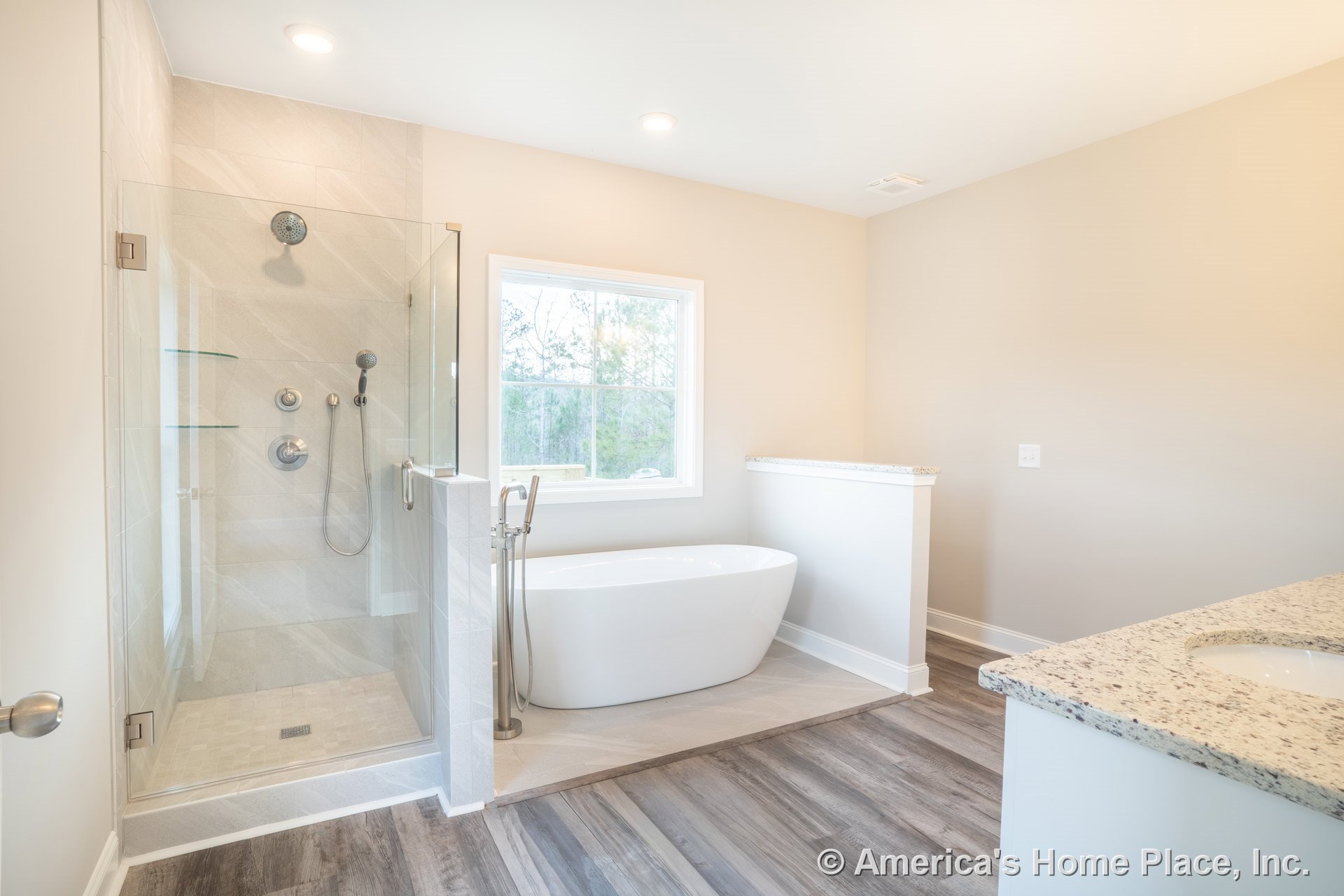 Frameless glass shower with tile walls, freestanding soaking tub, granite vanity countertop with white cabinetry, wood-look vinyl plank flooring, large window with decorative trim