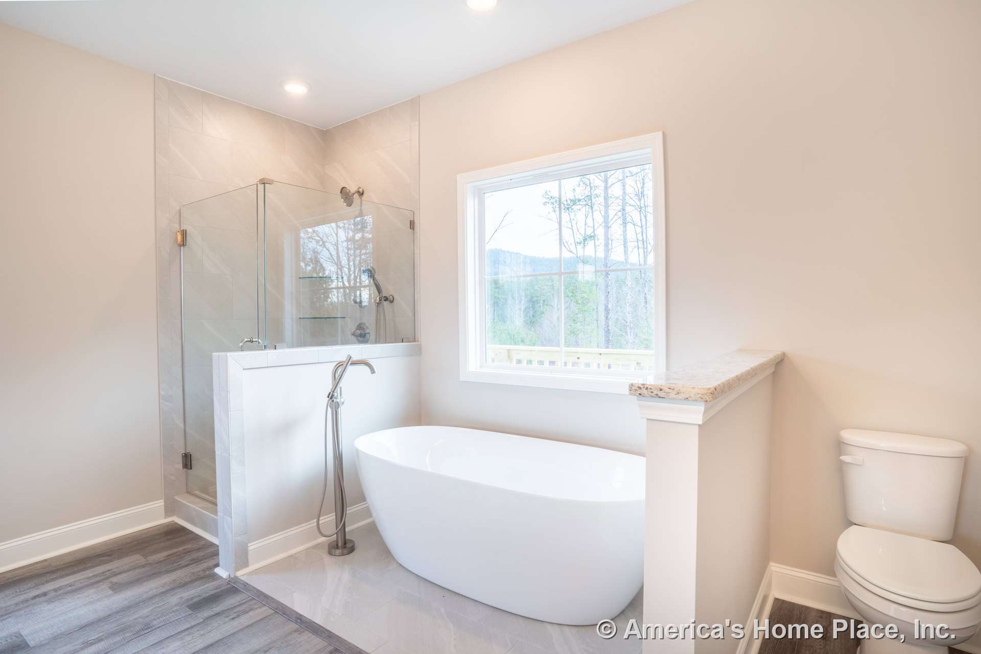 Freestanding soaking tub beside a granite-topped half wall, glass-enclosed shower, large window with white trim, wood-look tile flooring, recessed ceiling lights, and modern