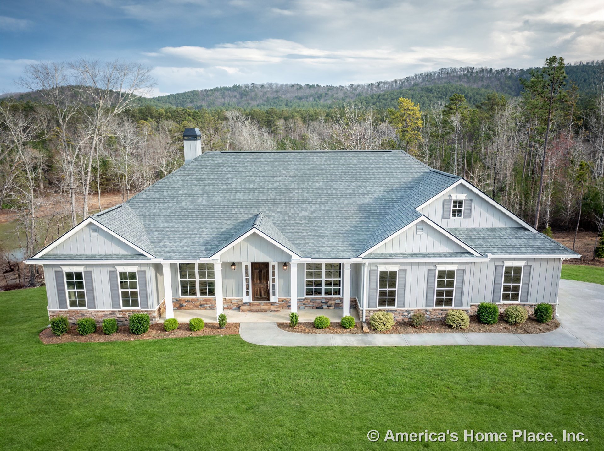 Board and batten siding with stone foundation trim, covered front porch supported by columns, multiple large front windows, gabled roof with chimney, concrete driveway, landscaped