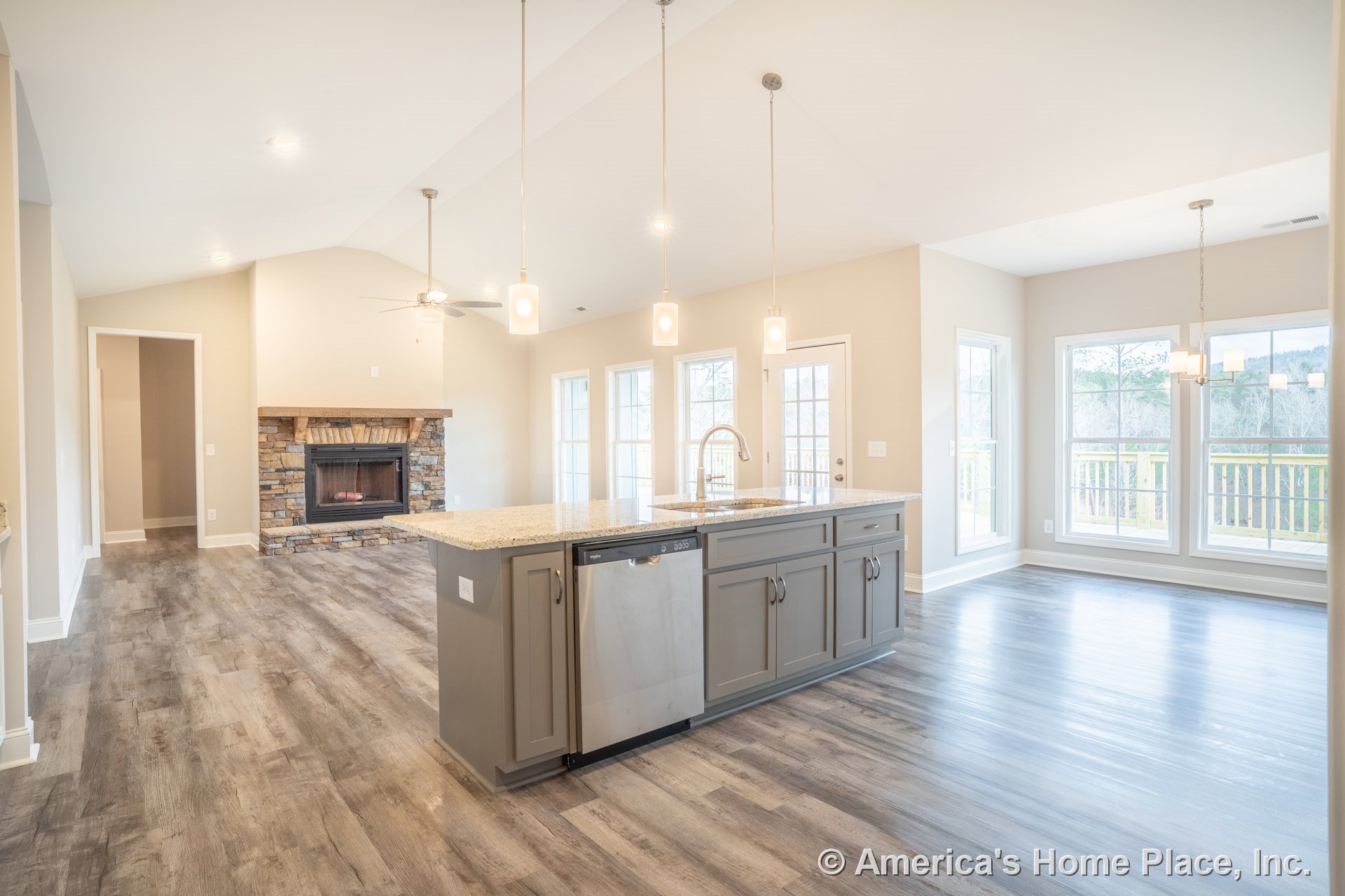 Open-concept living area with vaulted ceiling, recessed lighting, stone fireplace with wood mantel, wide plank wood-look flooring, multiple large windows, gray kitchen cabinetry