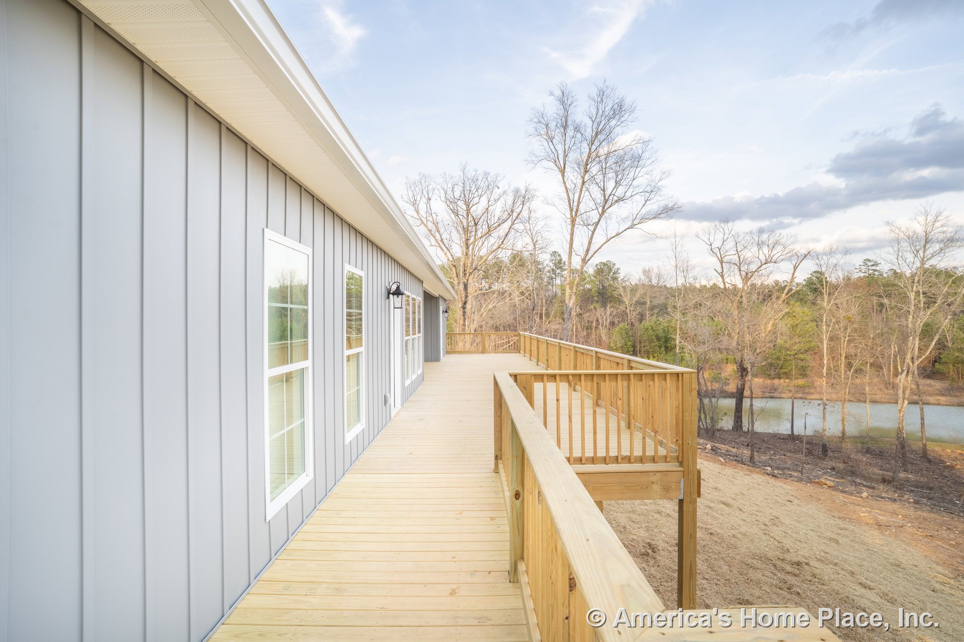 Spacious wooden deck with sturdy railing, vertical gray siding, multiple exterior windows, outdoor wall lighting, and overhanging soffit along the home's facade.