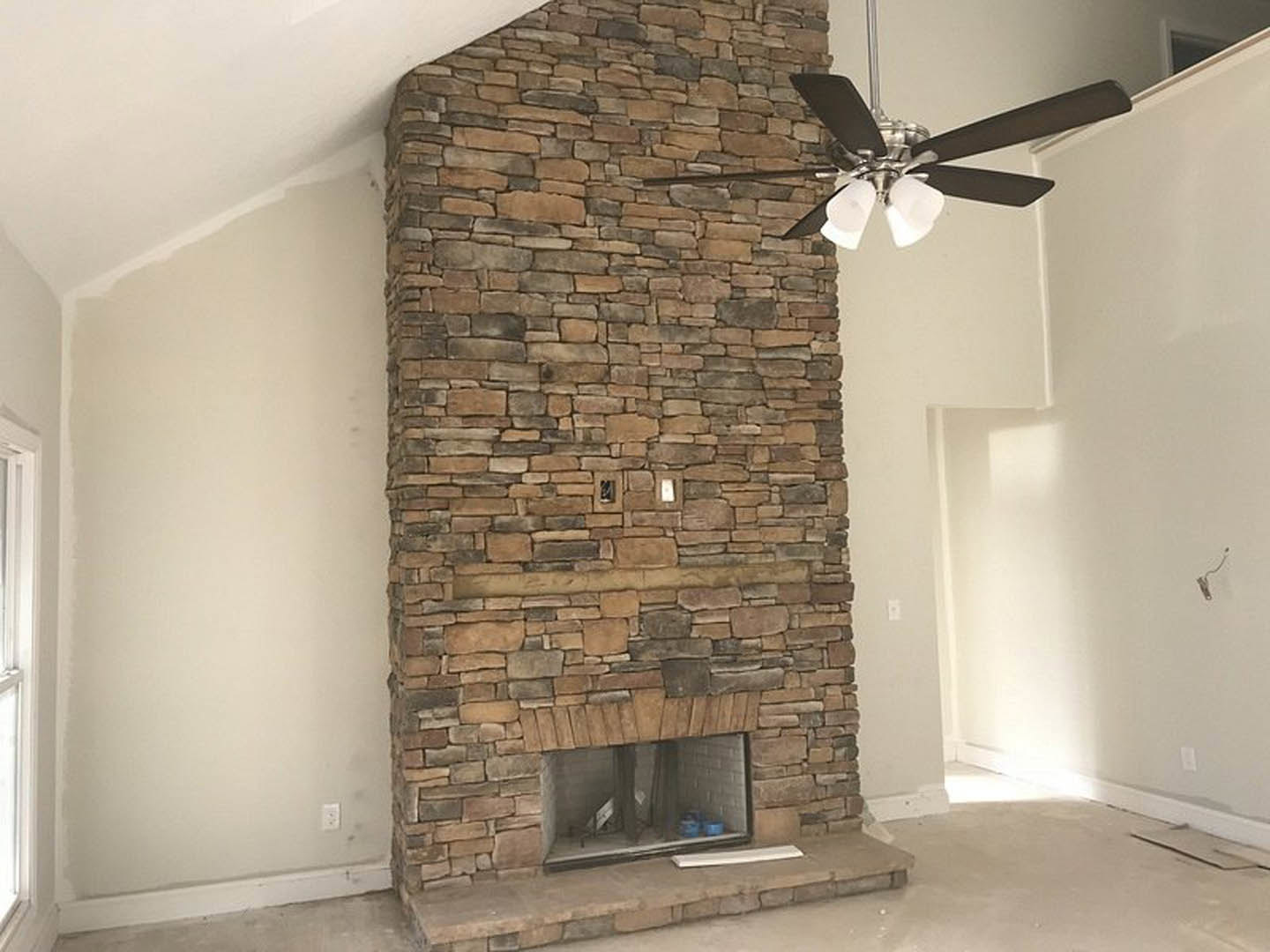 Ceiling fan with four blades above a living room featuring a brick fireplace, stone accent wall with light switch, wood-framed window, white walls, and a visible wire on a white