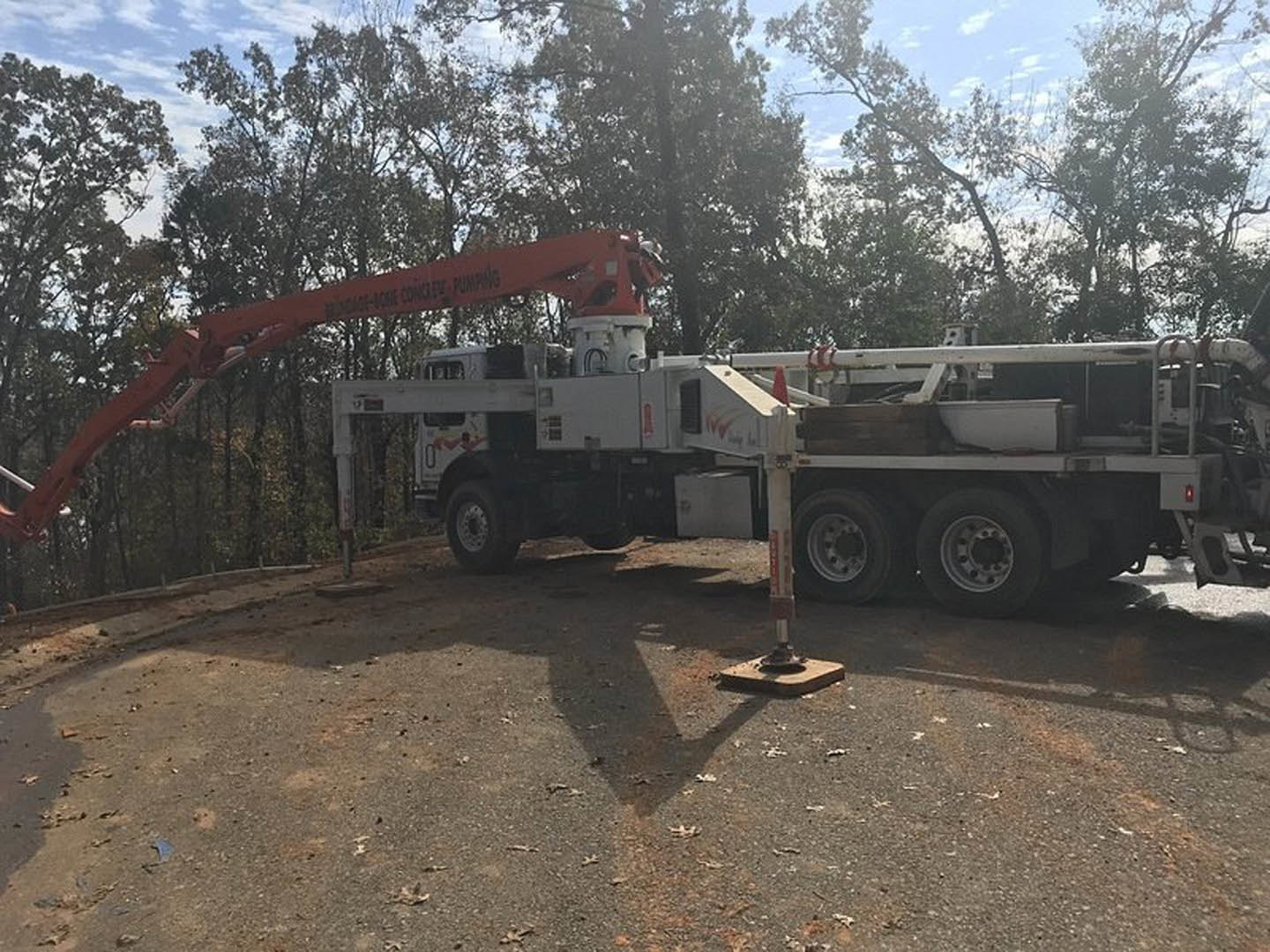 Large white truck with mounted crane parked on gravel driveway, tire and crane arm visible, leafy tree and blue sky in background