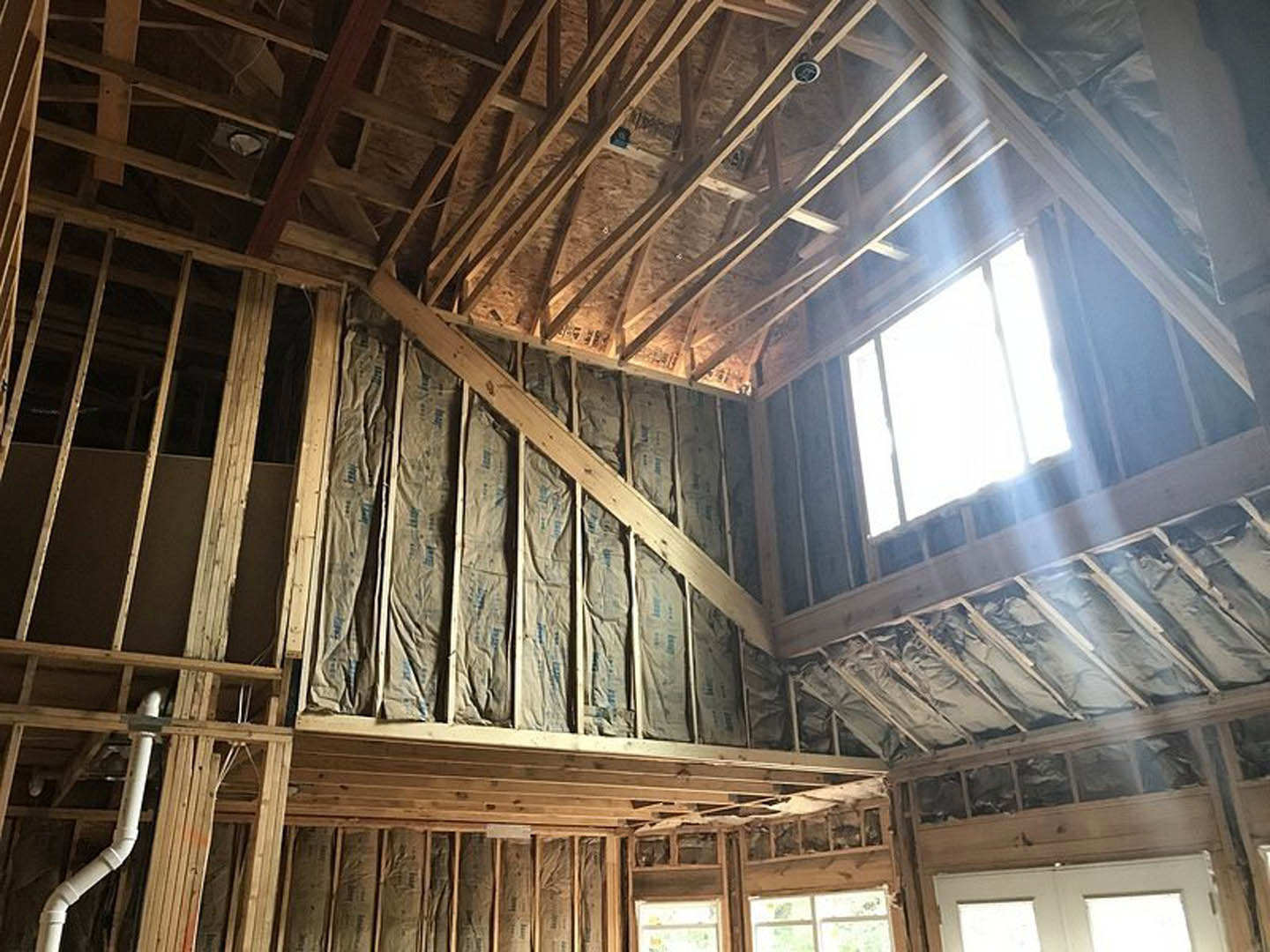 Wooden framing and exposed beams with wall insulation, white plumbing pipe, unfinished doorway, and sunlight streaming through window in house under construction