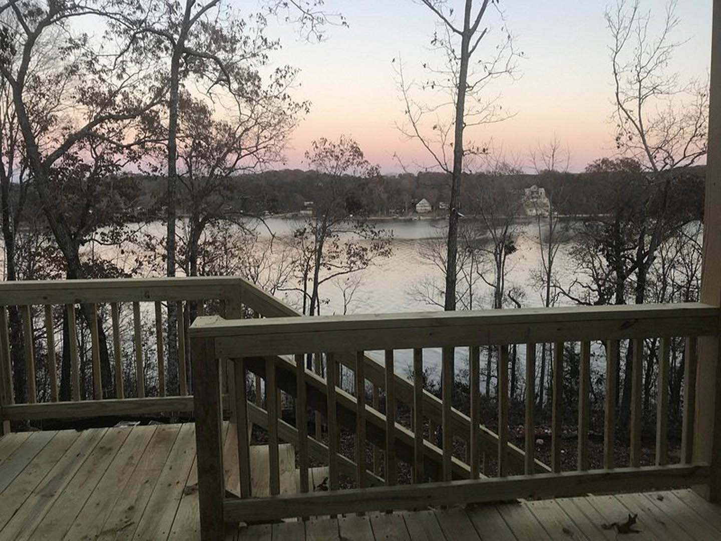 Wooden deck with railing overlooking a lake surrounded by trees and a pink sky