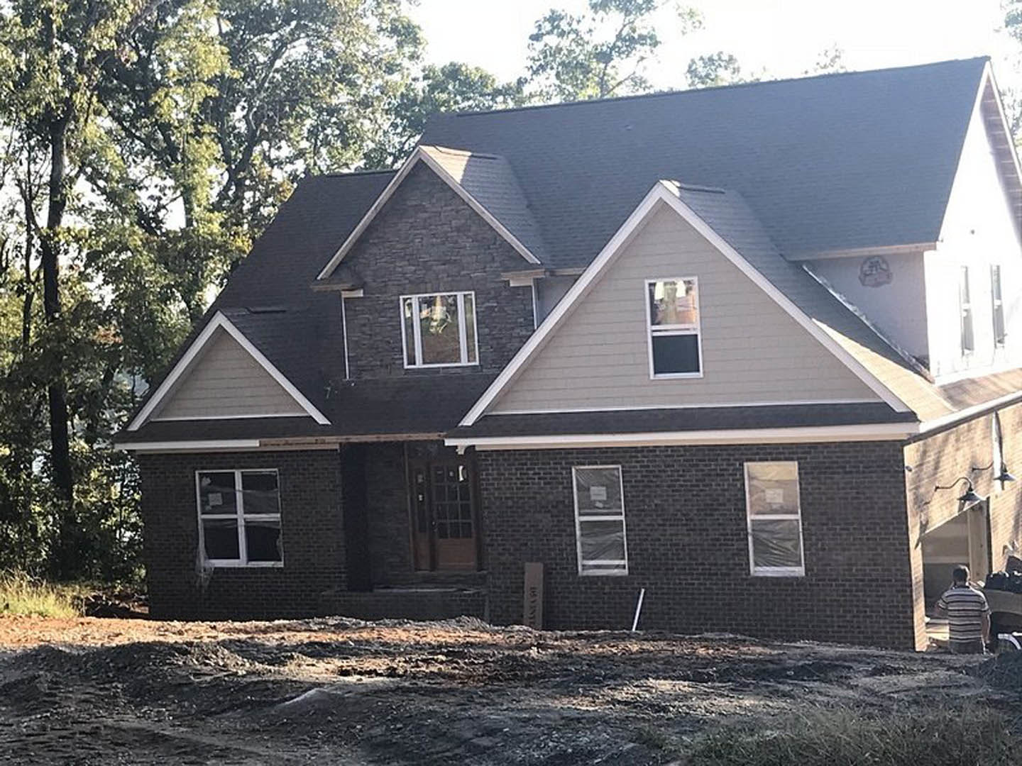 Partially built house with exposed framing, multiple white-framed windows, black board covering one window, man in striped shirt standing outside, trees in background