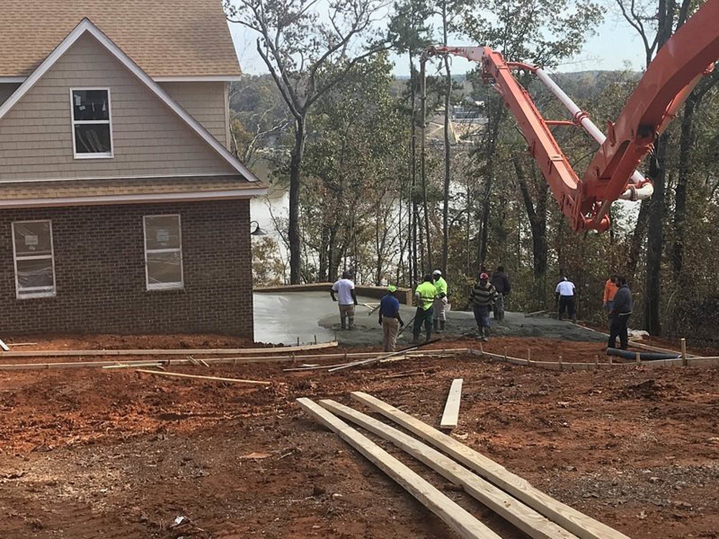 Several people standing on dirt near a partially constructed house with wood planks, white-framed window, and visible foundation; trees in background.