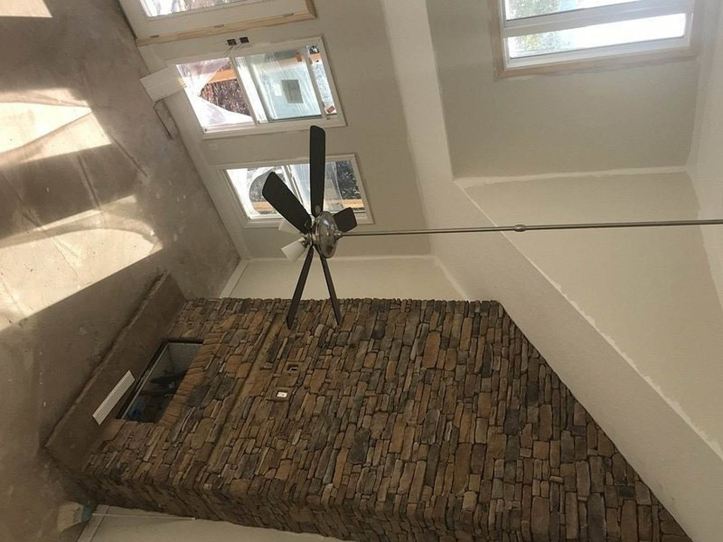 Ceiling fan mounted on exposed brick wall above hardwood floor, sunlight streaming through nearby window, casting shadows across interior space.