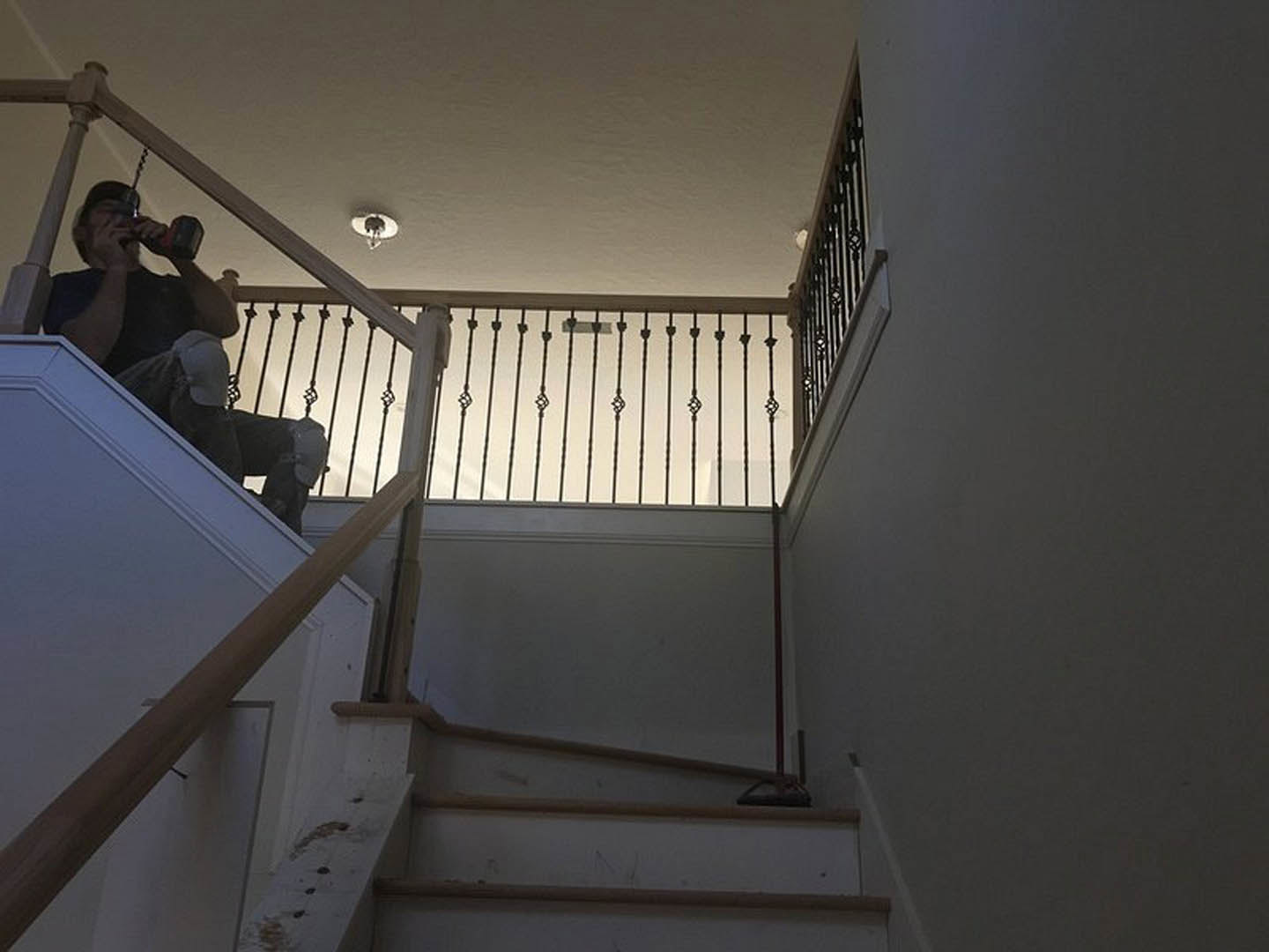 Wood staircase with metal balusters, plaster walls, and a man seated on a step holding a black object, possibly taking a selfie; close-up of modern light fixture visible.