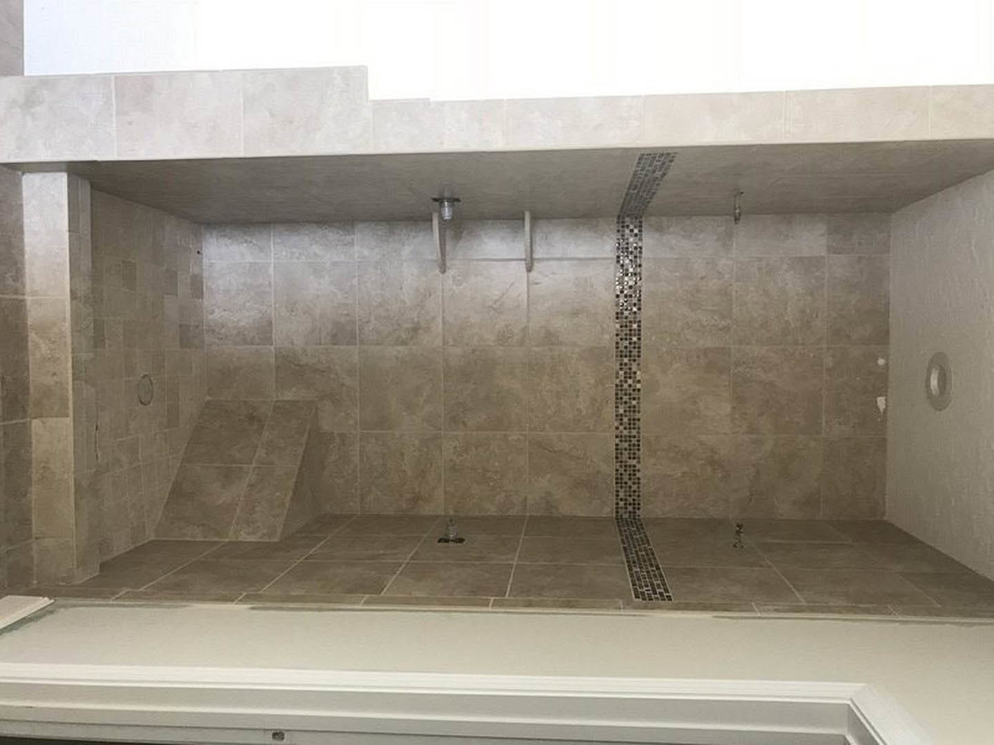 Shower with marble tile walls, white plaster ceiling with brown trim, circular white object and bottle on stone tile floor