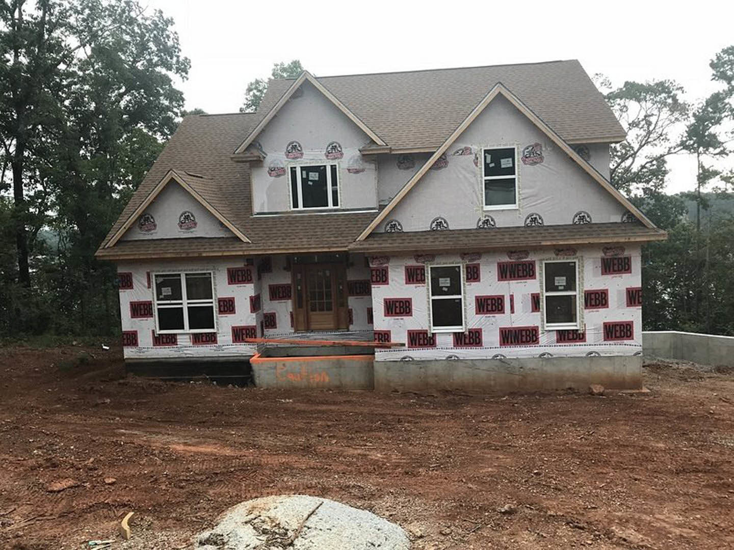 Partially built house with exposed framing, red and white construction signs attached to exterior walls, unfinished stonework, visible windows, and surrounding trees.