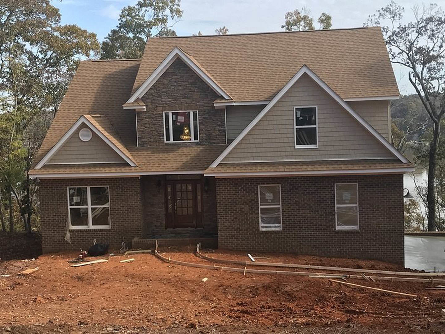 Two-story custom home under construction with brick and stone exterior, dirt yard, surrounding trees, white-framed windows, and unfinished entry door