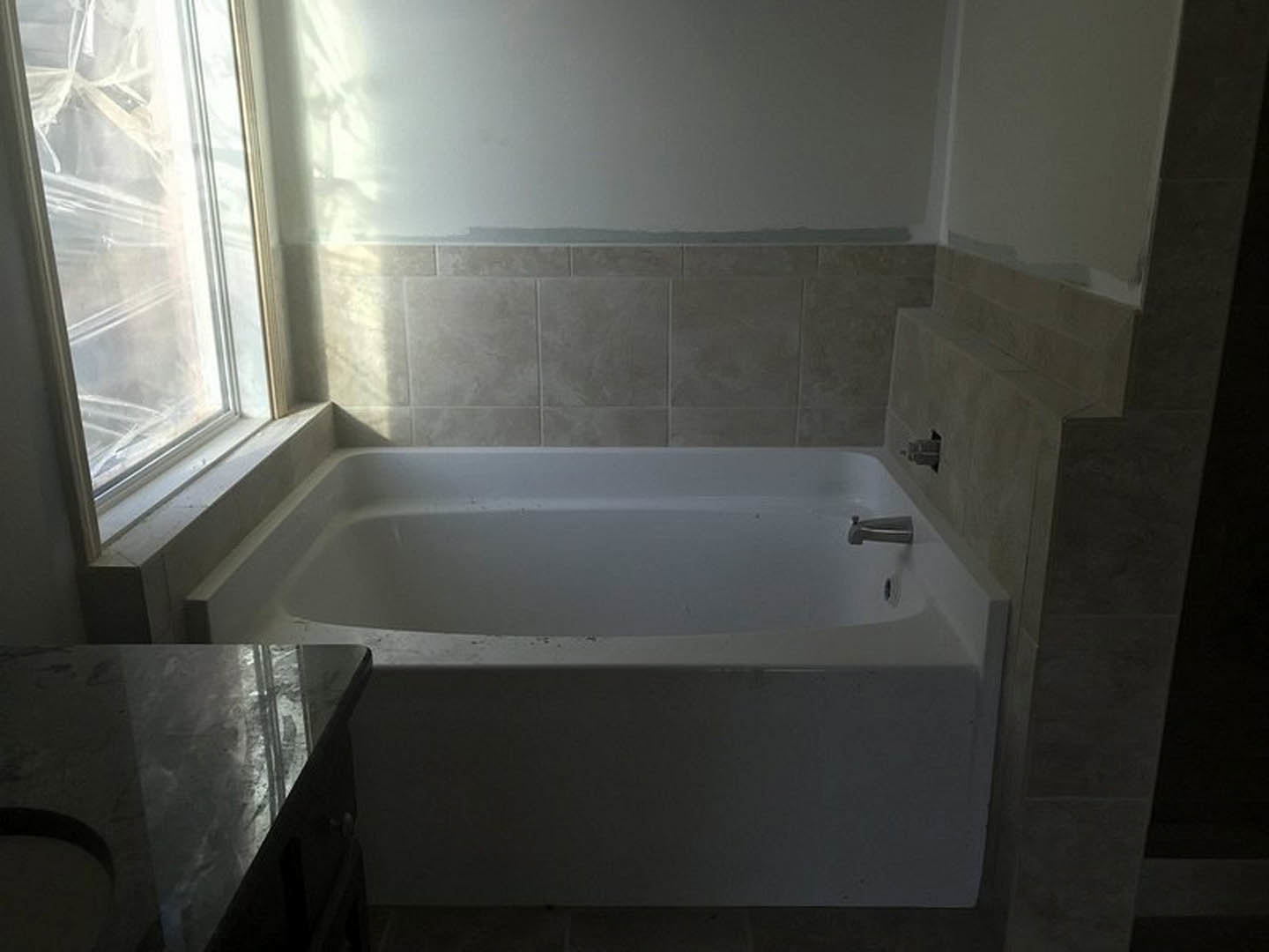 Freestanding white bathtub with chrome faucet, surrounded by light gray tile walls, large window, and modern bathroom accessories
