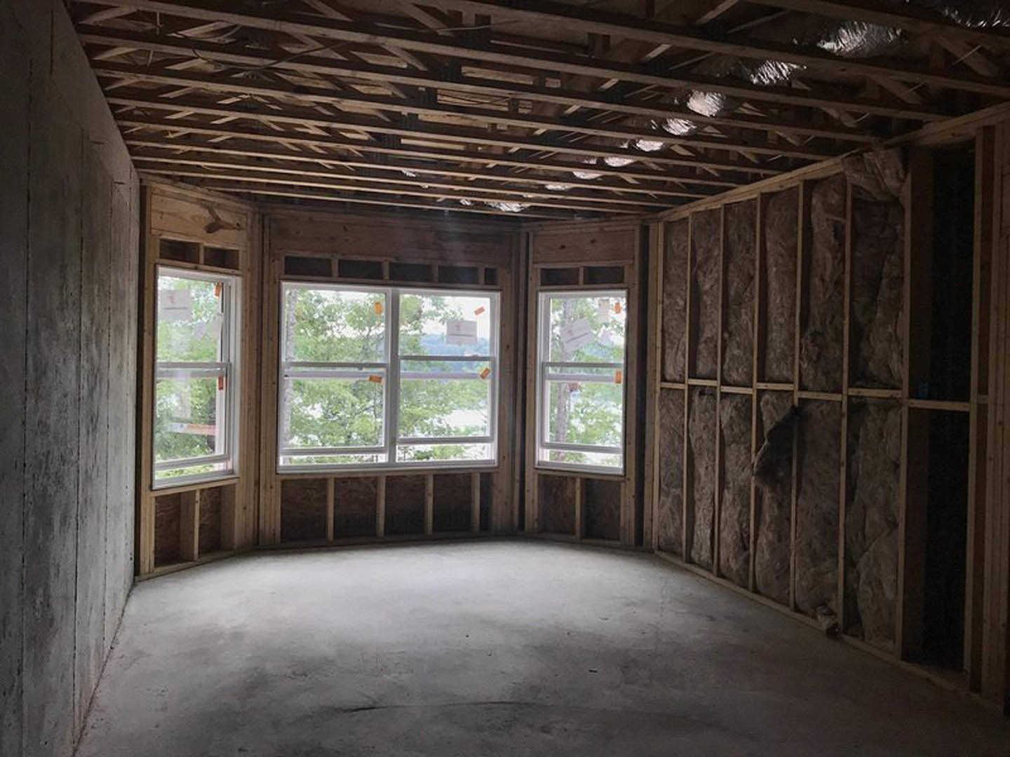 Spacious room featuring large windows with white frames, polished concrete floor illuminated by natural light, and wooden ceiling with exposed beams; view of trees visible through