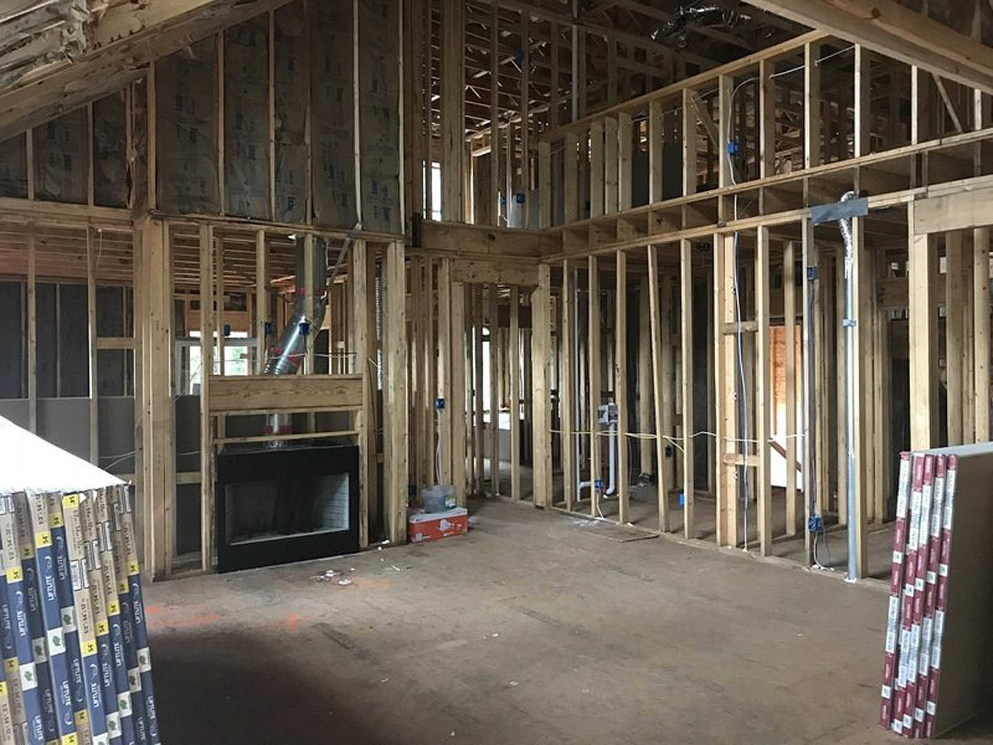Living room with exposed wood framing, modern black fireplace with glass front, built-in shelves holding stacks of books, light-colored flooring, and unfinished walls.