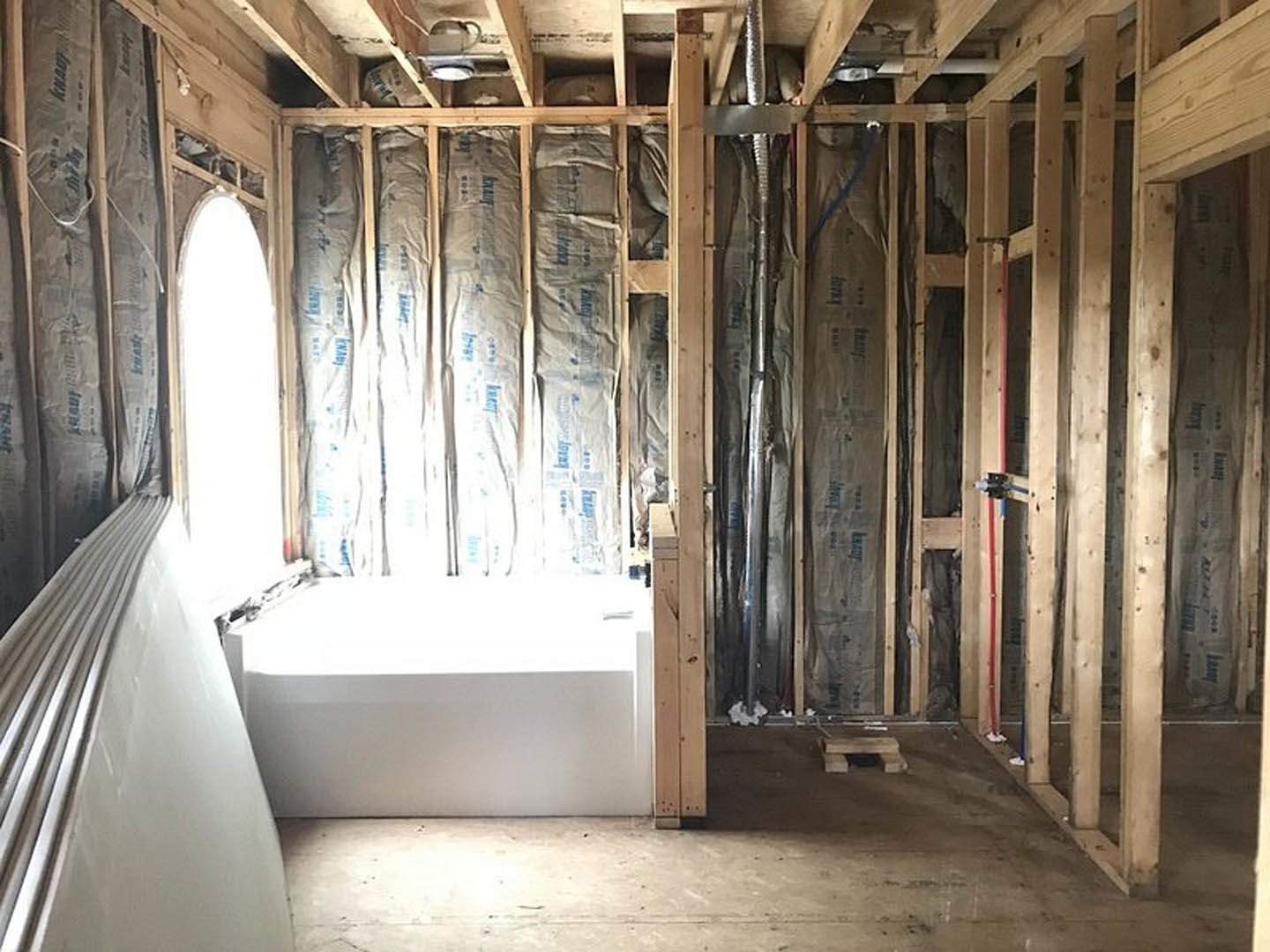 Bathroom under construction with exposed wood beams, unfinished plaster walls, white rectangular bathtub, and construction materials scattered on the floor.