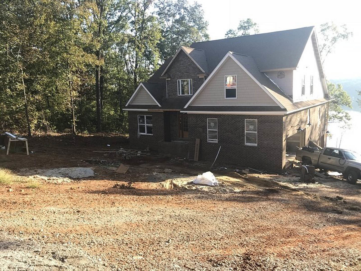Wood-framed house under construction surrounded by tall trees, exposed plywood walls, construction materials scattered on bare ground, partially installed windows, white object