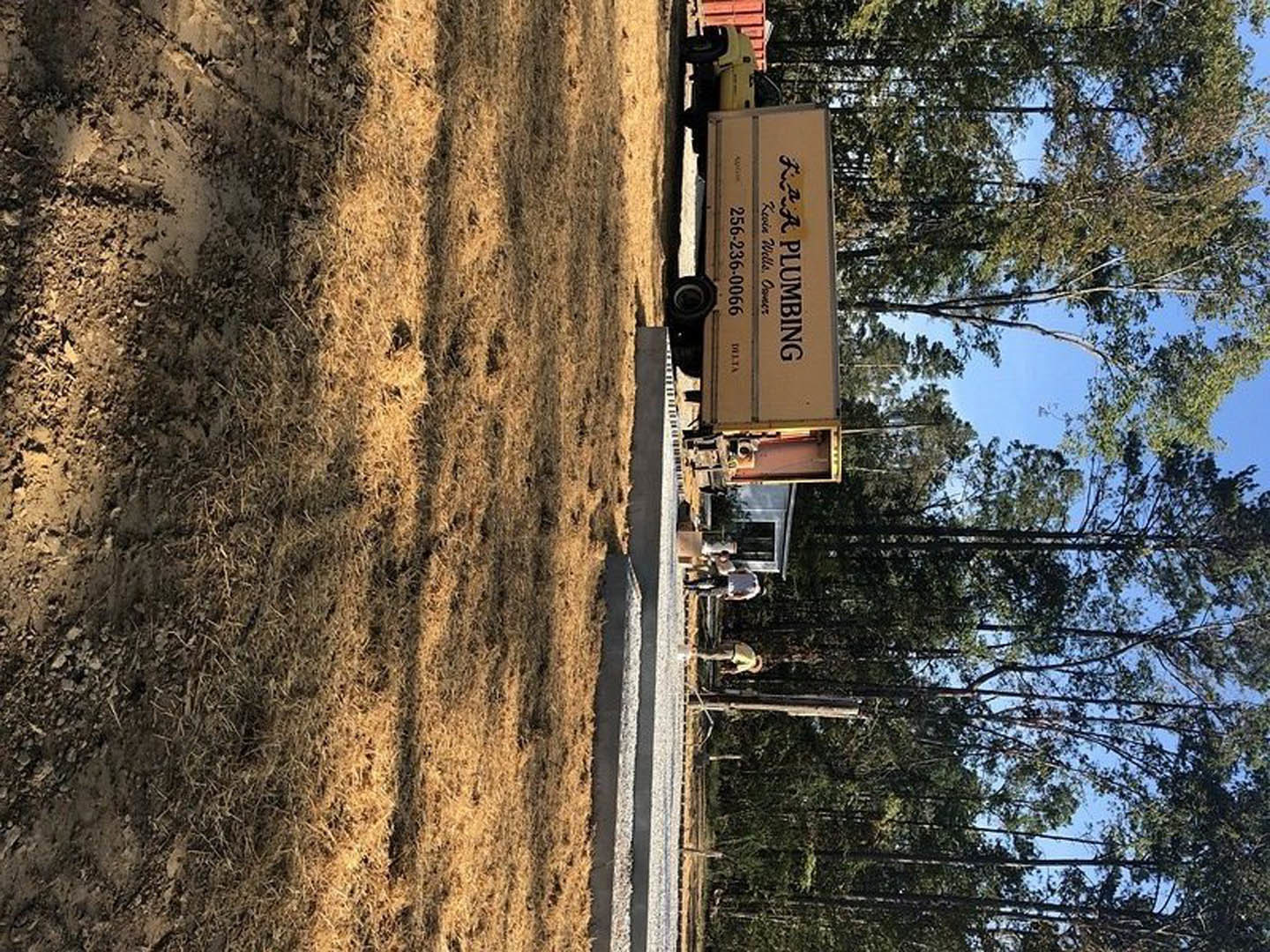 White pickup truck parked on dirt and grass field beside a group of trees under blue sky, black text sign visible in background, several people standing near tree line.