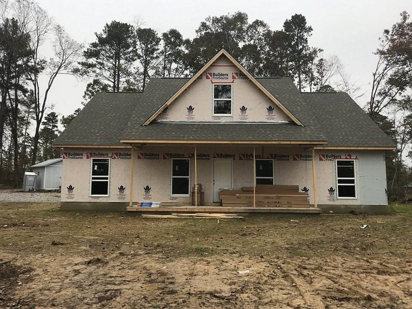Framed wooden house under construction on a dirt lot, surrounded by trees, exposed beams and plywood visible, clear sky overhead