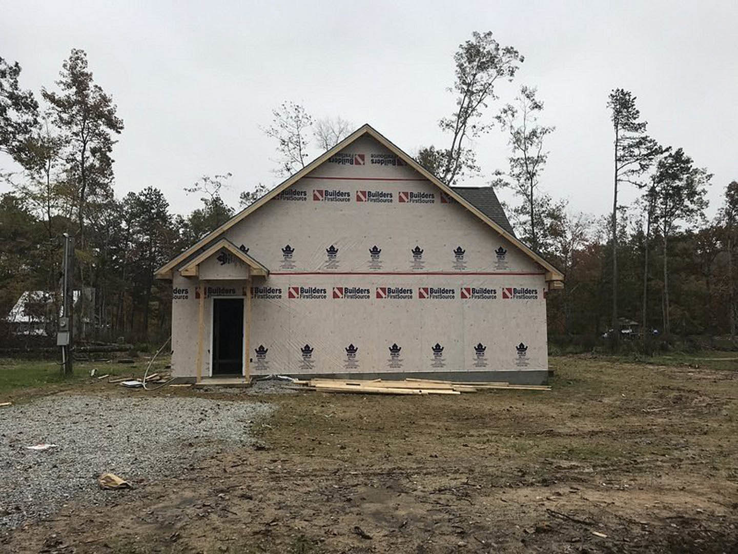 Partially built house with exposed insulation panels, shingled roof, piles of gravel and wood on dirt lot, surrounded by mature trees.