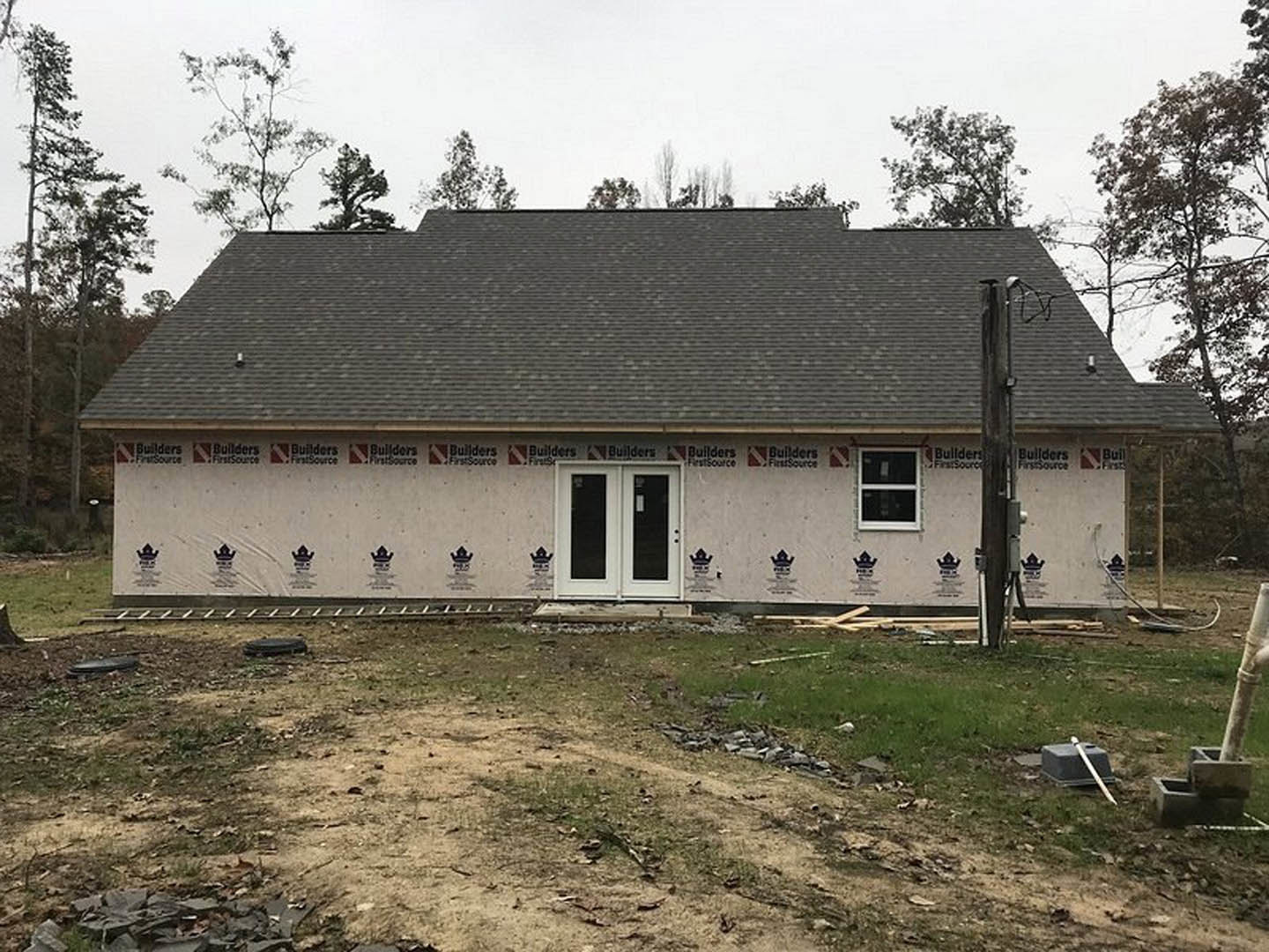 Modern house under construction with white framed windows, double glass doors, unfinished yard with patches of dirt and grass, surrounded by trees and open sky