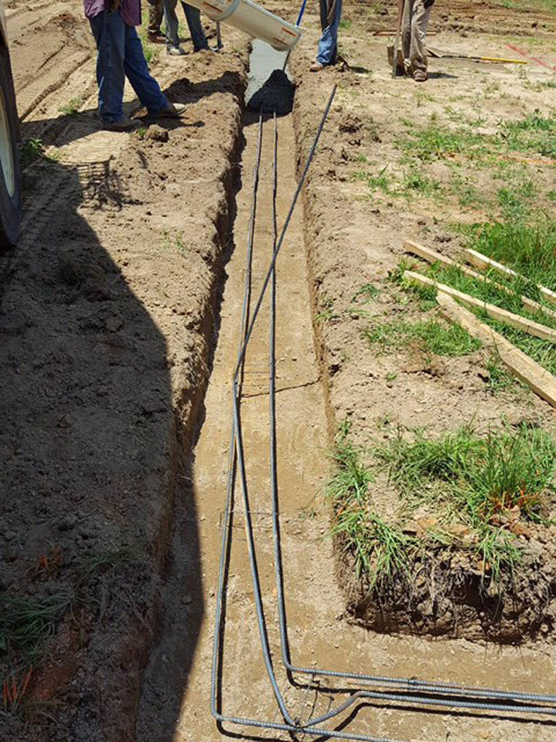Man wearing work boots standing on dirt beside open trench with exposed black metal pipes, surrounded by soil and sparse grass.