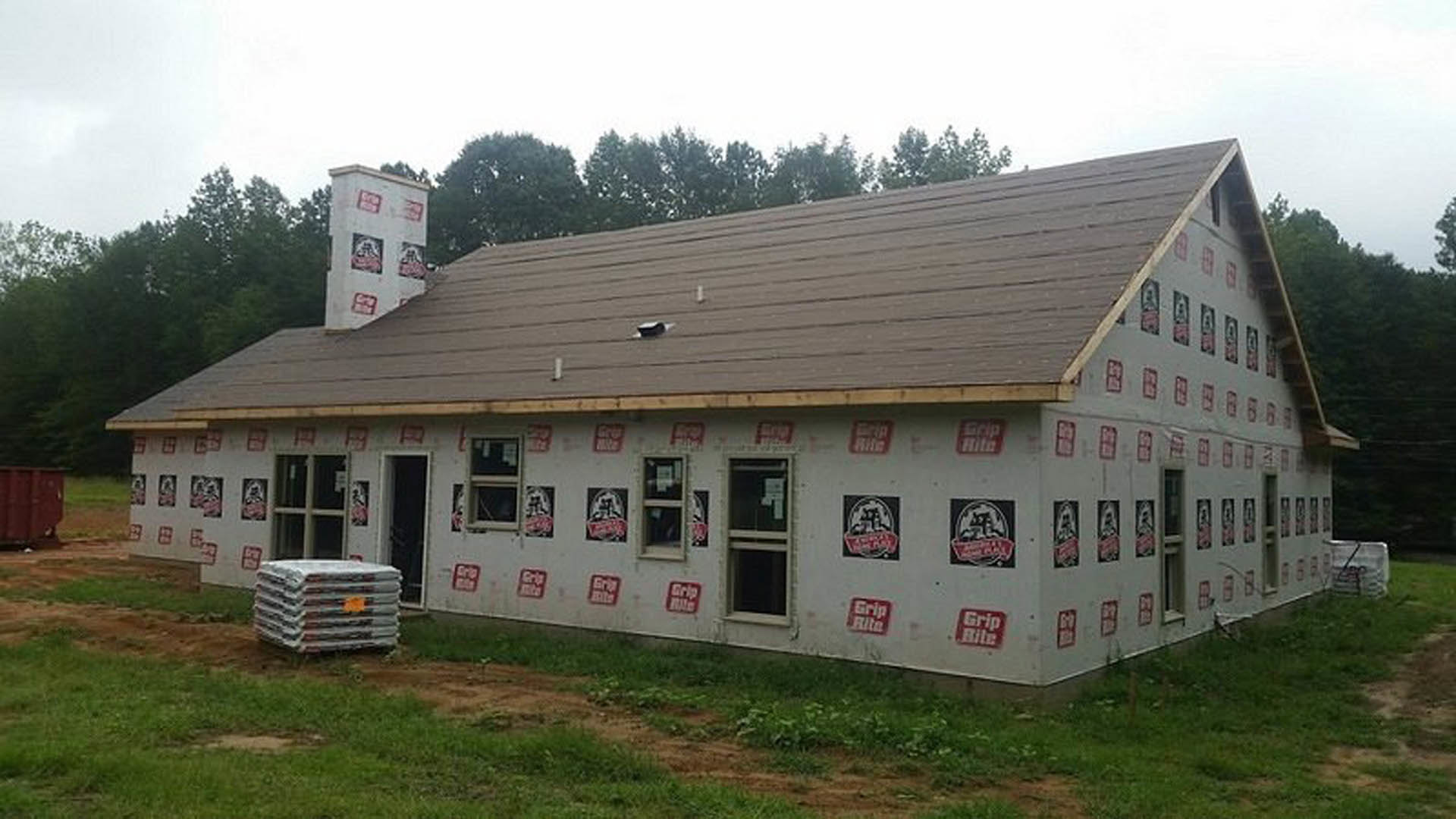 Partially built house with exposed sheathing covered in branded stickers, visible window with numbered sticker, pitched roof, grassy lot, and trees in background
