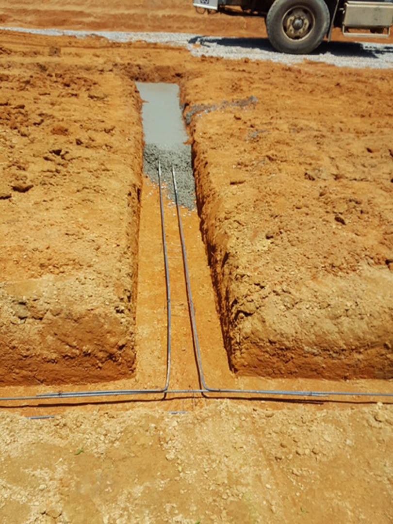 Trench dug in soil with exposed metal pipes, construction truck tire visible nearby