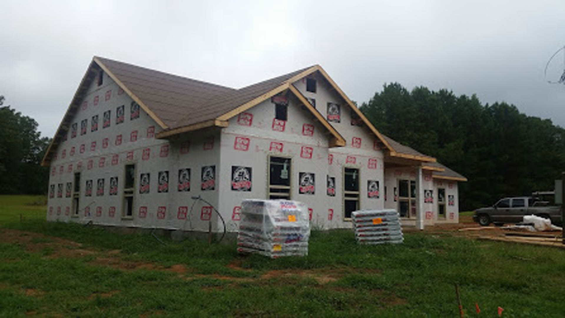 Modern house with light-colored siding, pallets of construction materials stacked along the side, grassy lawn, scattered trees, and a parked car partially visible in the