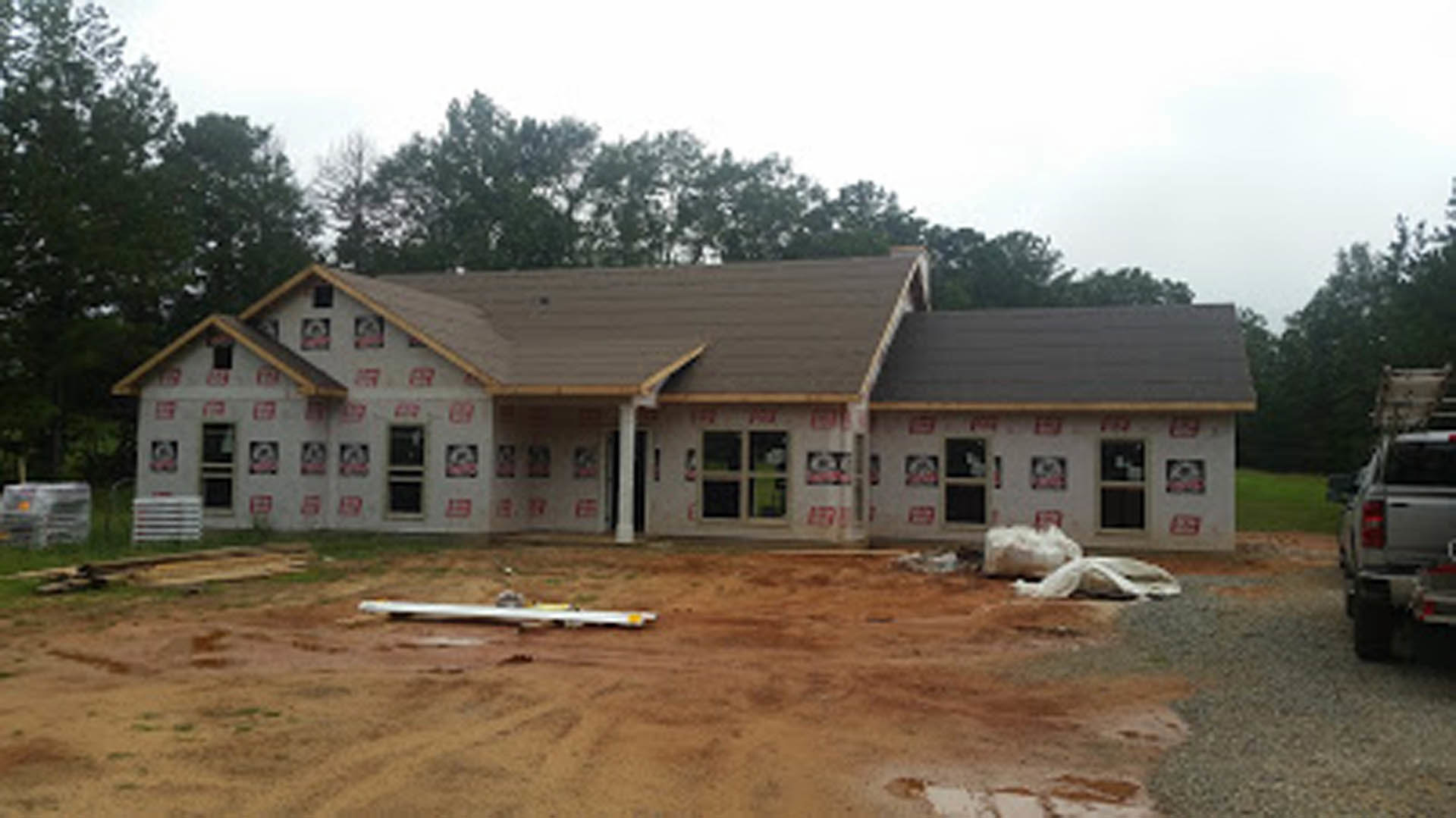 Framed house under construction with exposed roof trusses, surrounded by trees, construction truck parked in front