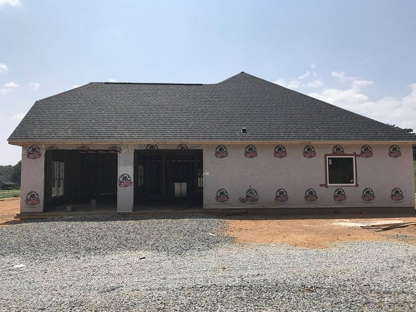 Two-story house under construction with exposed framing, gravel driveway, garage door, and windows beneath a cloudy sky