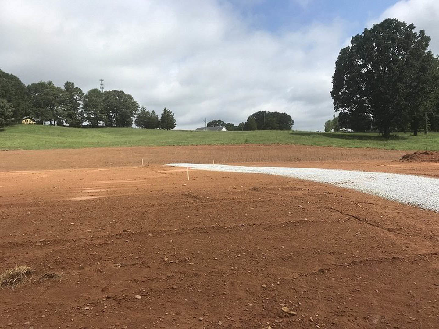 Dirt field with a white line, large leafy tree, blue sky with scattered clouds, blurry building in background, light pole near tree