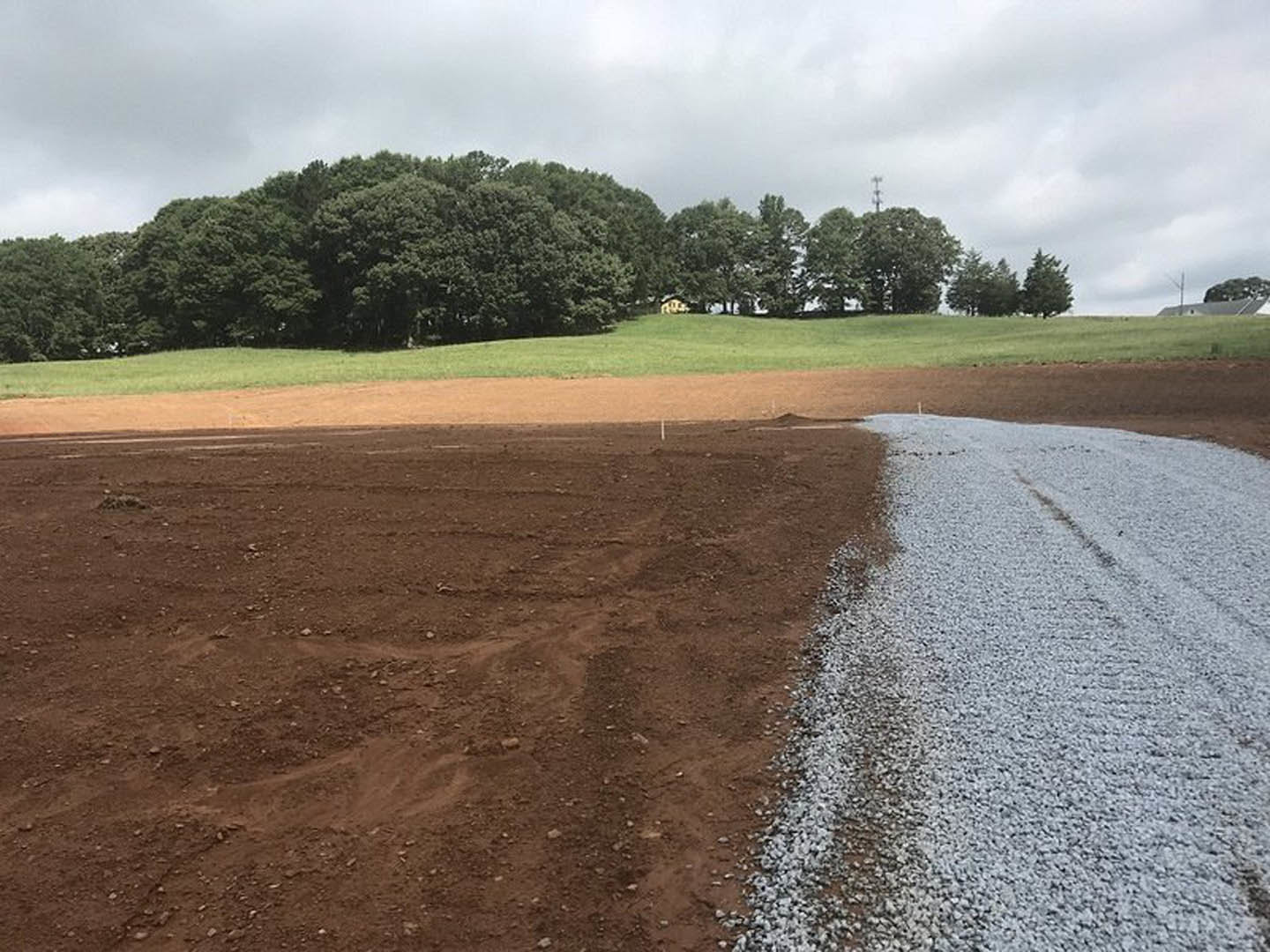 Gravel road with tire tracks leading to a custom home set in a grassy field, surrounded by trees under a partly cloudy sky, white pole visible near dirt area