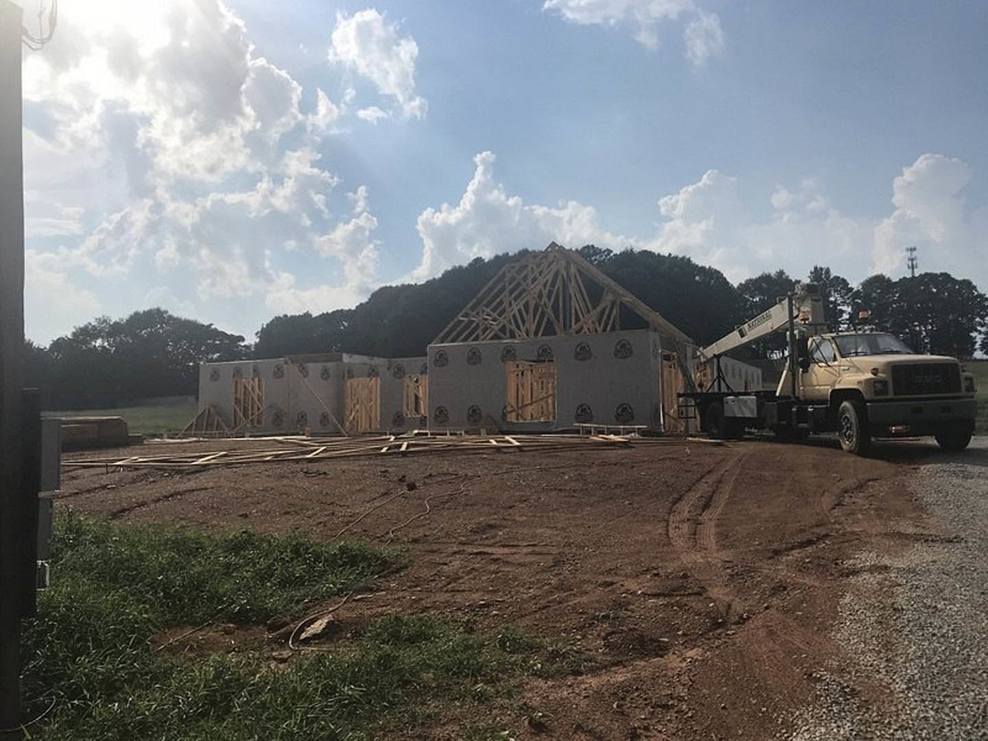 Wood-framed house under construction with exposed beams, dirt lot, construction truck parked beside structure, cloudy sky overhead, trees in background
