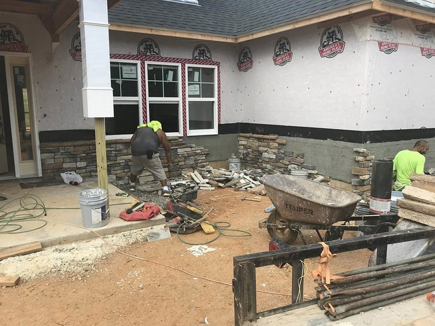Man in green shirt moving lumber and wheelbarrow near house under construction, exterior ground covered with building materials and tools