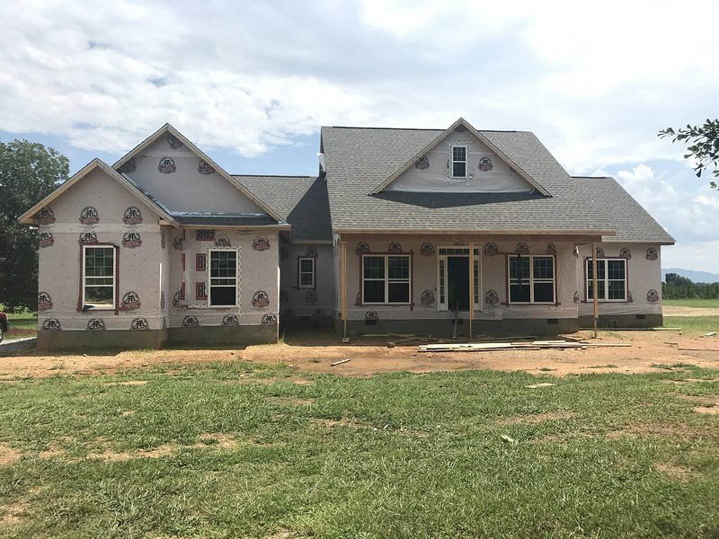 Two-story house under construction with white-framed windows, partially finished exterior walls, and a yard featuring grass and exposed dirt patches.