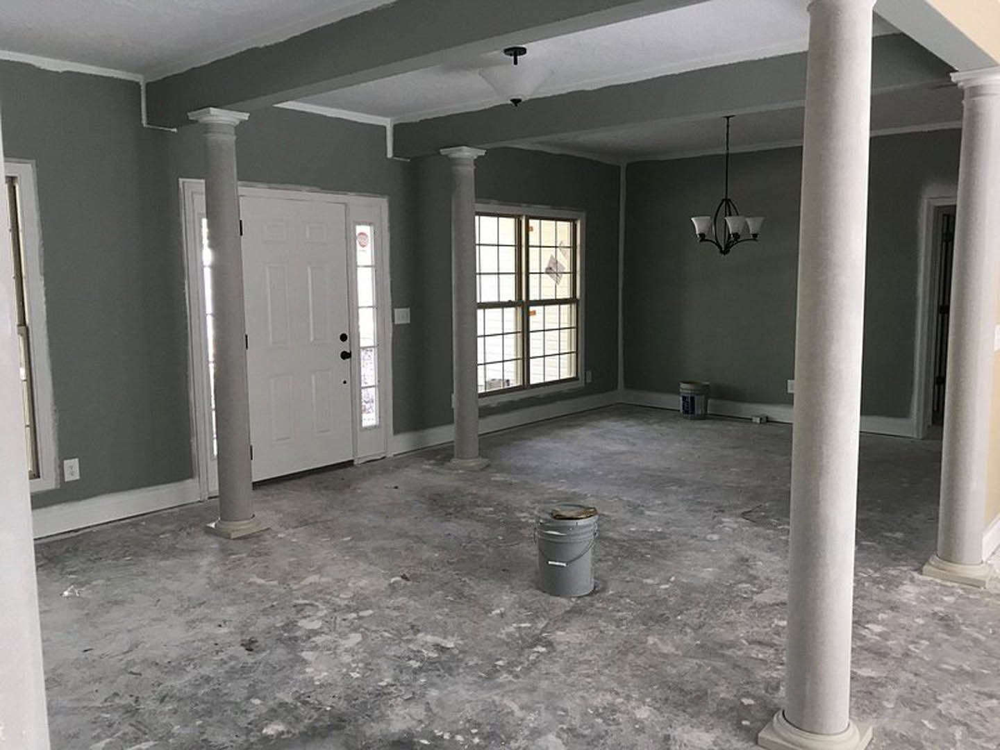 Open living space with white pillars, dark wood door, polished tile floor, modern chandelier, window with trim, grey bucket with wooden lid, and paper towels on a countertop