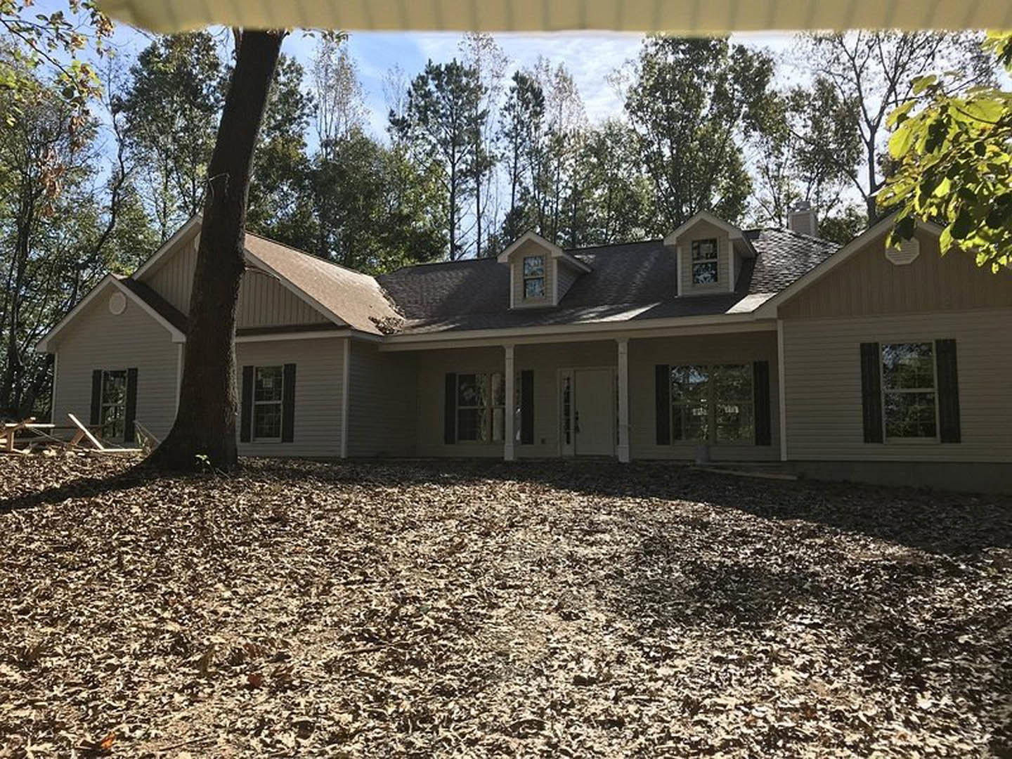 Two-story cottage-style home with white-framed windows, gray roof featuring a dormer, surrounded by mature trees and fallen leaves covering the ground