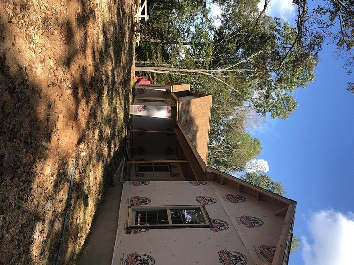 Framed house under construction with exposed wooden beams, large windows, dirt path in foreground, surrounded by leafy trees beneath a clear blue sky