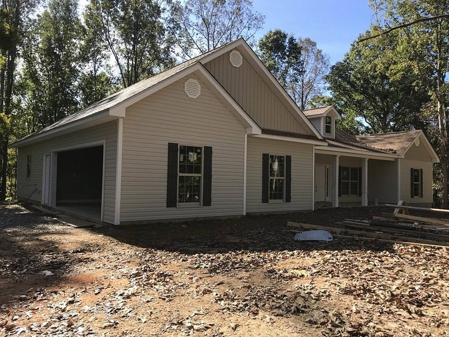 Partially built house with exposed framing, white window frame, wall vent, shingled roof, scattered leaves on ground, and mature trees in background