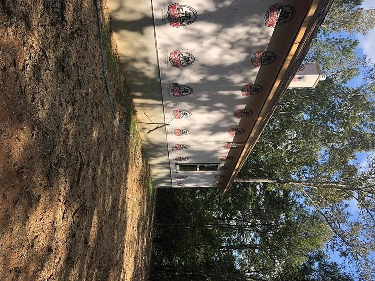 Exterior view of a house under construction with one wall covered in white insulation panels featuring red and black logos, dirt yard in foreground, hose on ground, trees and blue