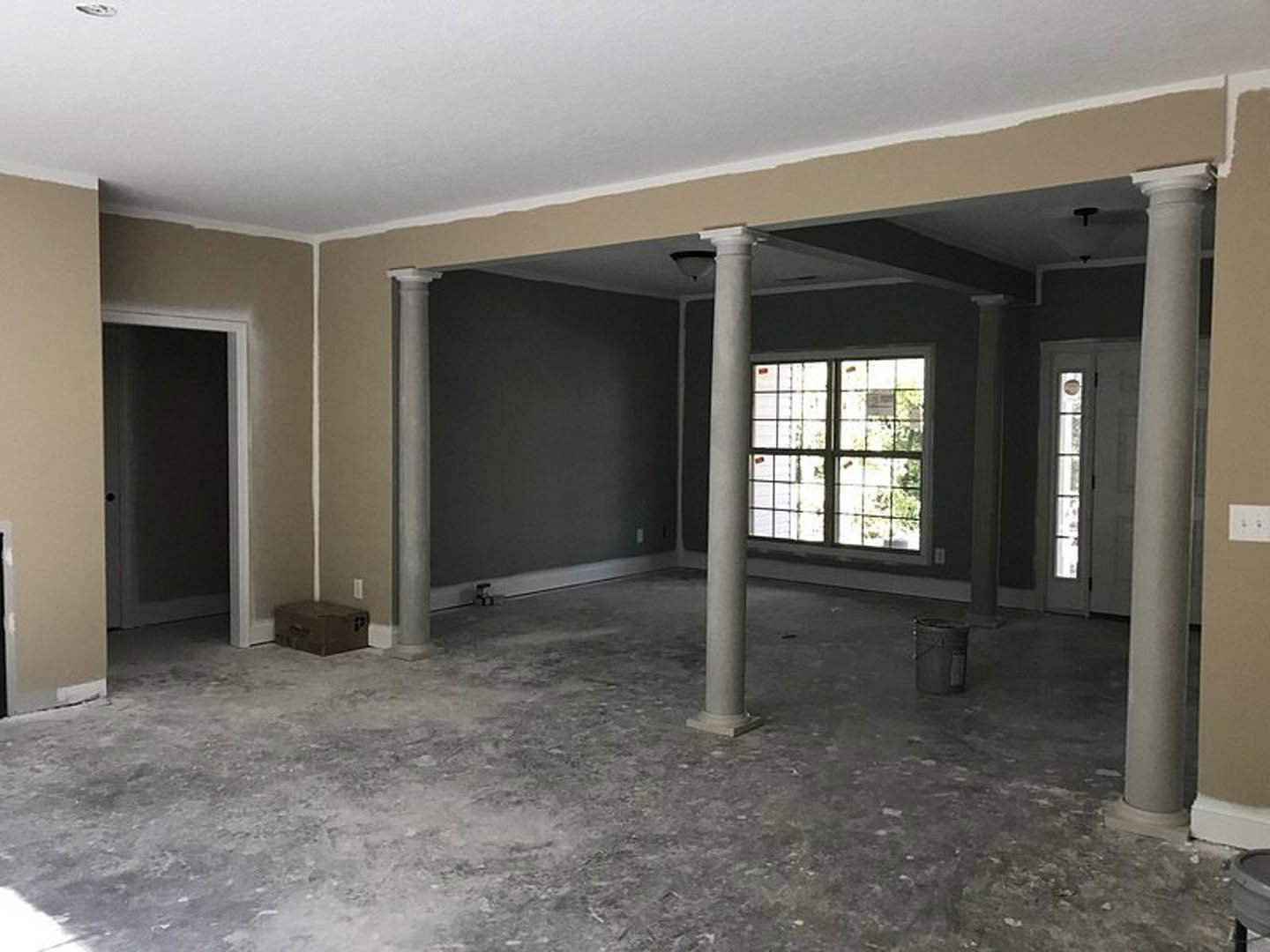 Open living space with white plaster columns, large window with natural light, neutral walls, light wood flooring, and modern door.