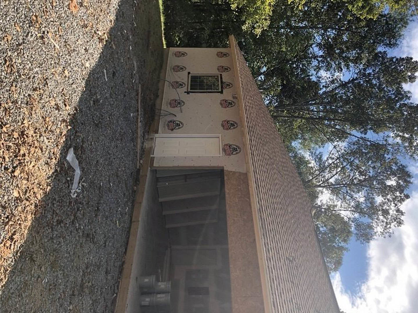 Partially built home with exposed framing, white window frames, temporary construction screens, and unfinished staircase; surrounded by trees and cloudy sky, white paper scattered
