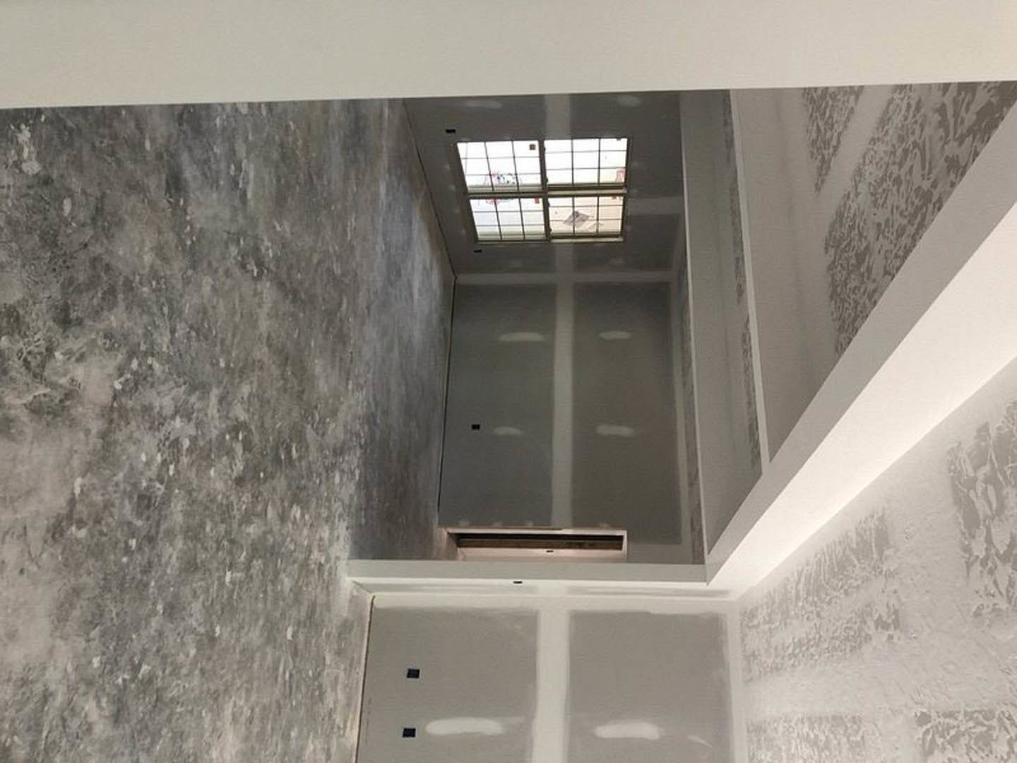 White plaster walls, large grid-style window, exposed concrete floor, flat ceiling, natural daylight filling minimalist interior.