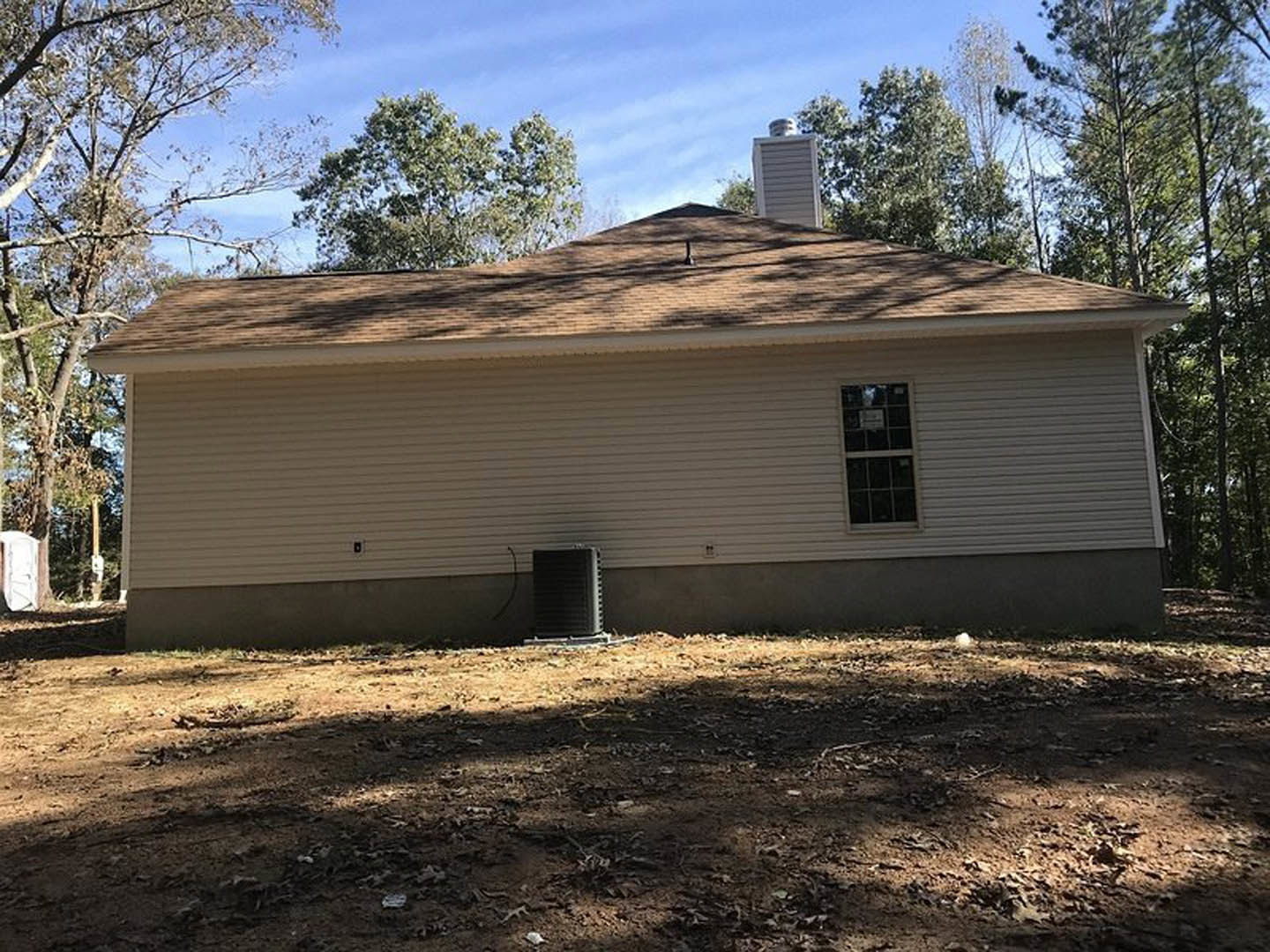 Two-story house with gray siding, brick chimney, black water tank beside exterior wall, large windows including one with a sign, shingled roof, dirt yard scattered with leaves